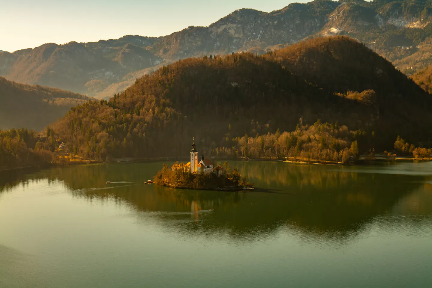 Lake Bled's island church in warm low light, surrounded by calm water and hills in late-autumn colour