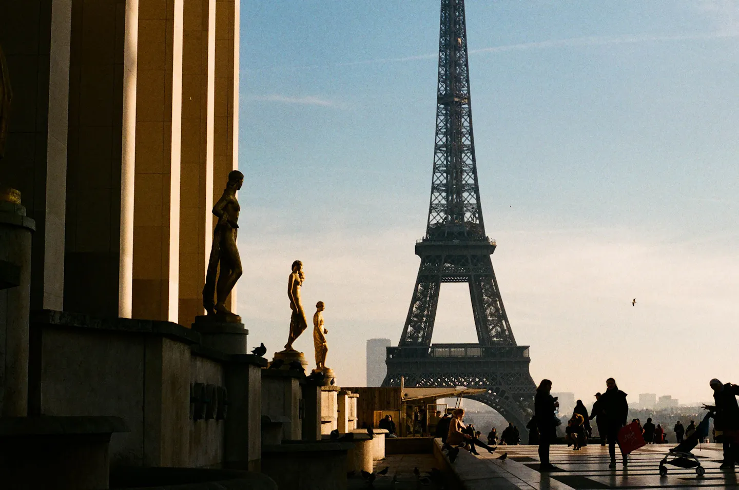 Gilded statues along the Palais de Chaillot terrace at Trocadero, backlit by low sun, with the Eiffel Tower standing across the river behind them