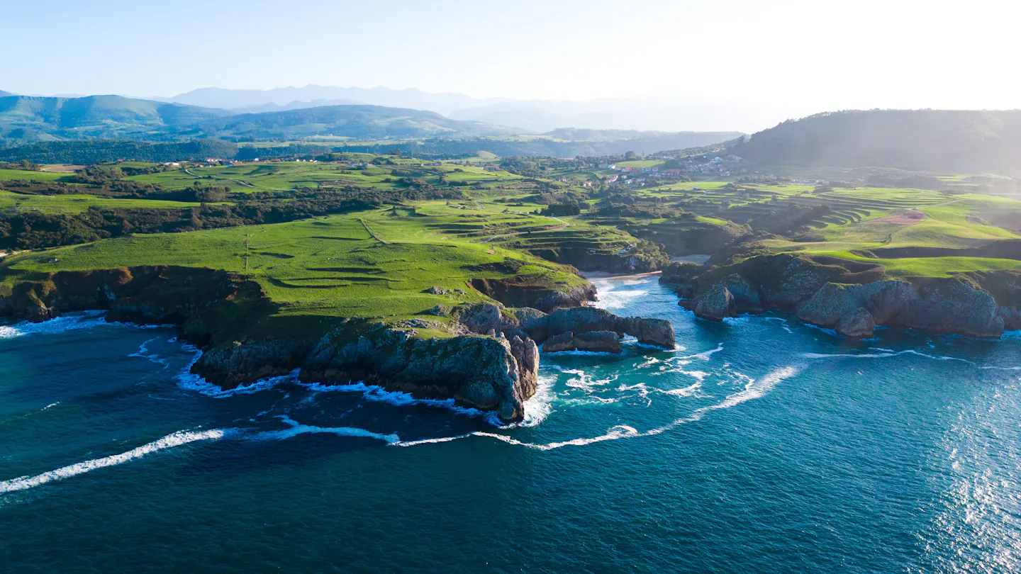 Green clifftop meadows and low headlands meeting the sea on the Cantabrian coast of northern Spain, with white surf breaking below