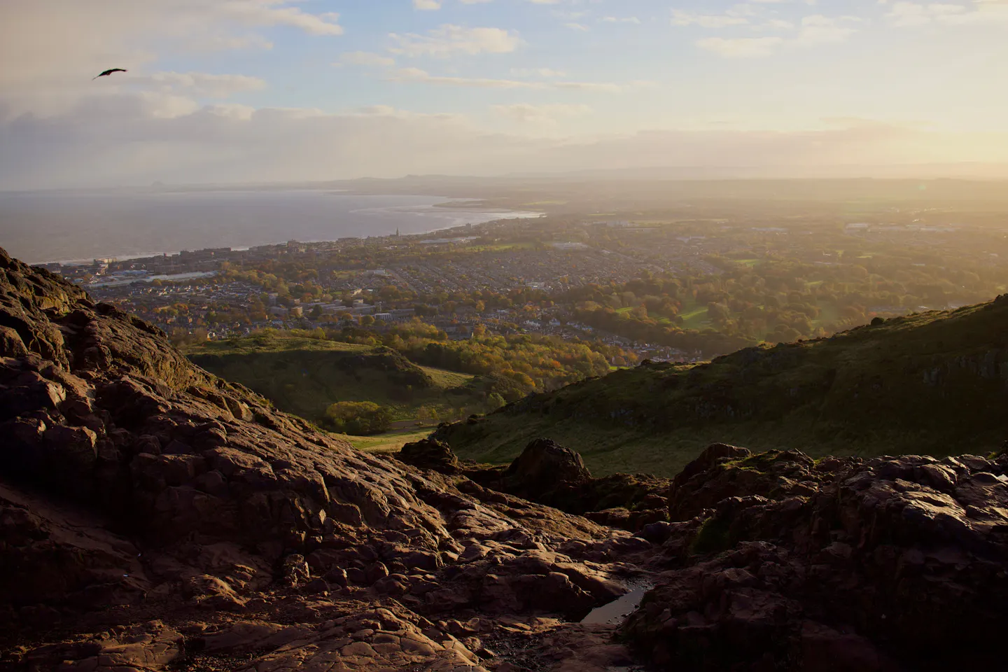 The view from the rocks of Arthur's Seat at golden hour, low sun raking across the city, autumn treetops and green hollows below, and the Firth of Forth stretching to the horizon