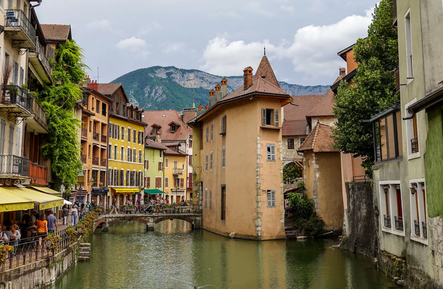 The Thiou canal in Annecy old town with the turreted Palais de l'Isle, ochre and yellow shuttered houses, a small stone footbridge, café awnings and an Alpine ridge behind