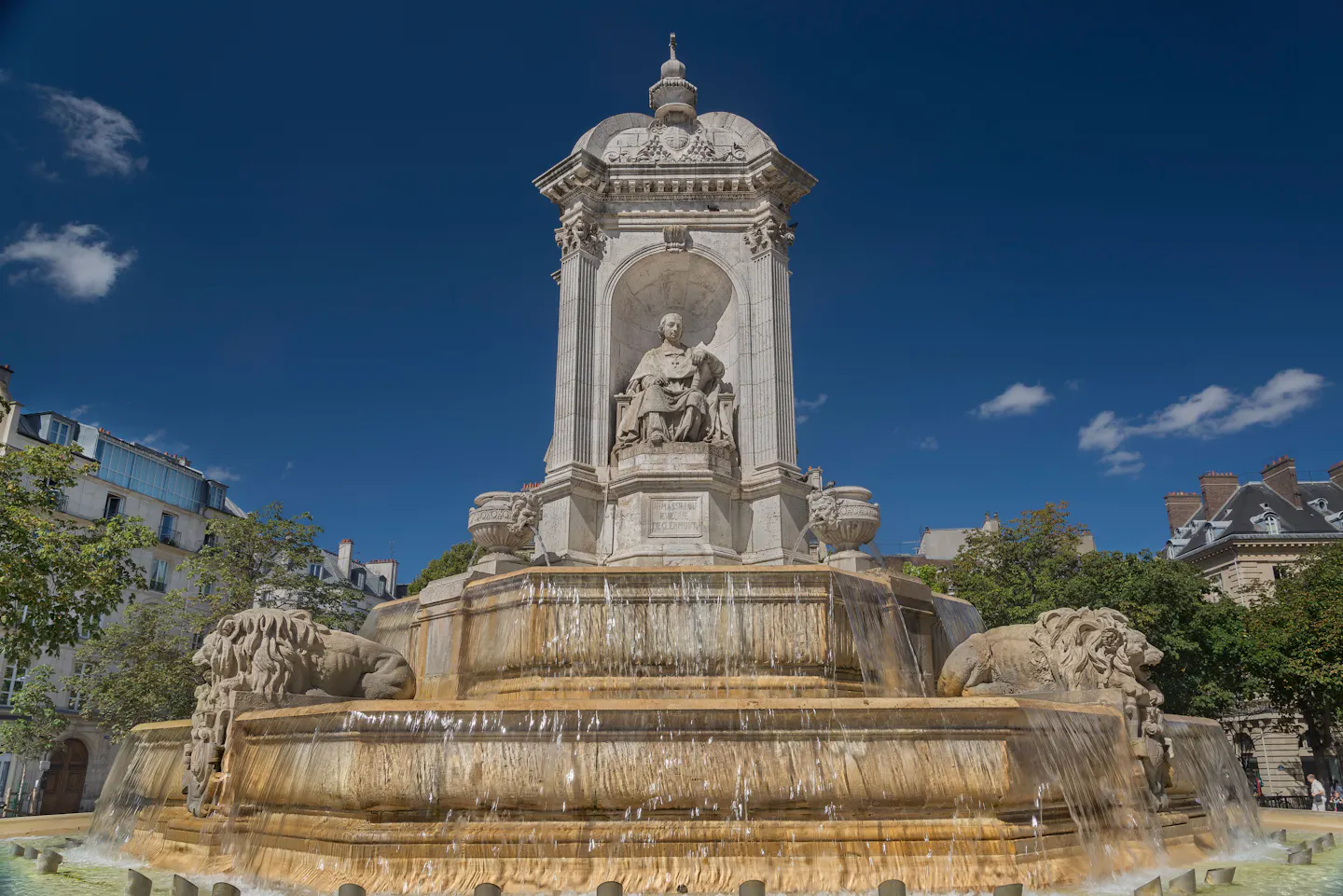 Water falling over the tiers of the Fontaine Saint-Sulpice, with its stone lions, under a deep blue sky