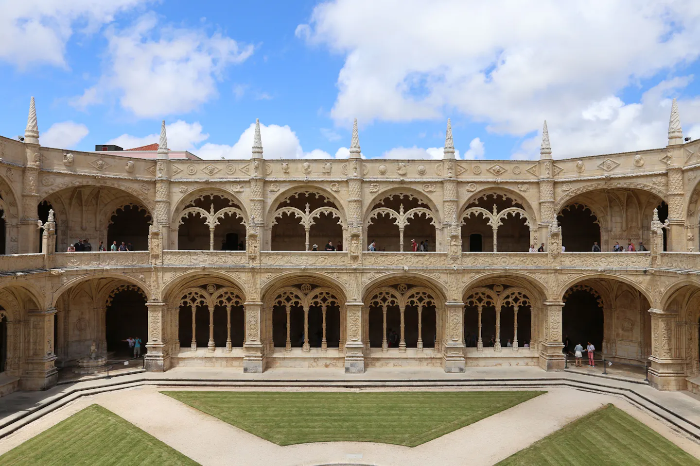 Two tiers of carved Manueline arcades running around the sunlit golden-stone cloister of the Jeronimos Monastery, under a blue sky with white clouds