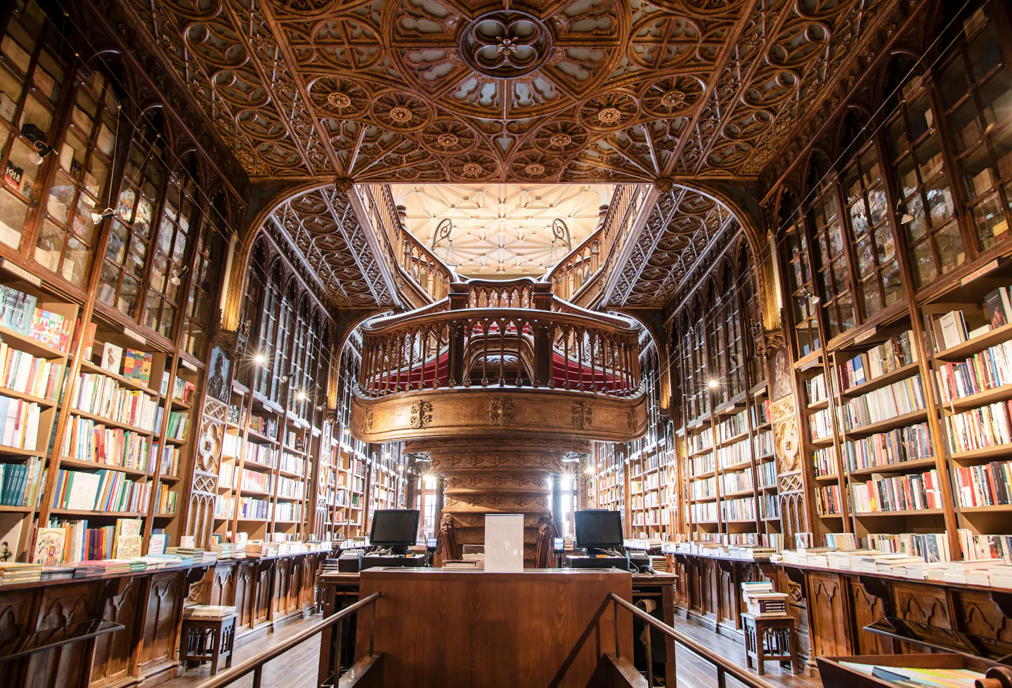 The upper gallery of Livraria Lello in Porto, carved wooden ceiling panels and Gothic-arched bookcases packed with books, looking down onto the famous curving red staircase