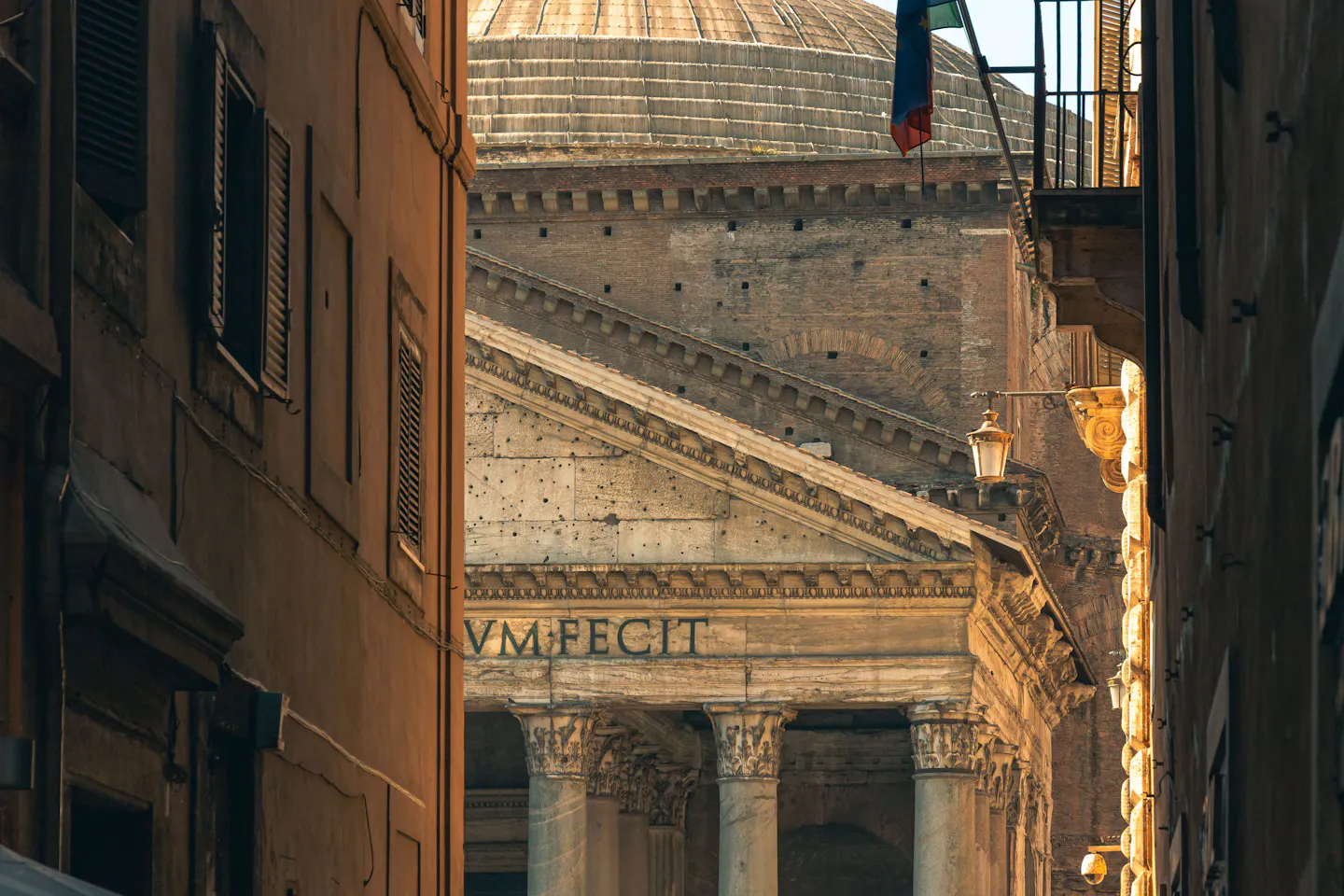 The Pantheon's columned portico and coffered dome glimpsed at the end of a narrow Roman alley at dusk, framed between warm ochre house walls
