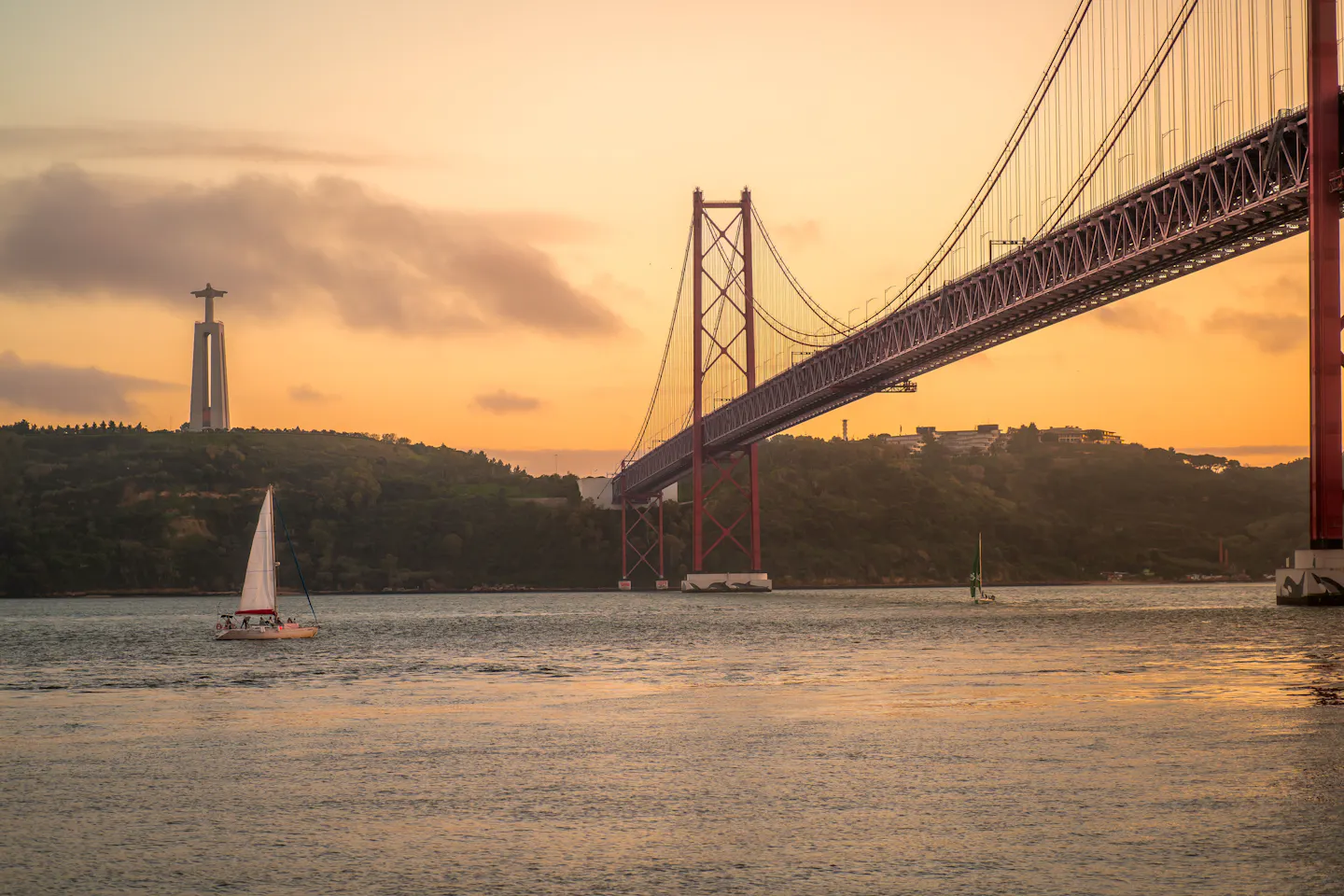 The red 25 de Abril bridge over the Tagus at golden hour, with a sailing boat and the Cristo Rei statue on the far bank