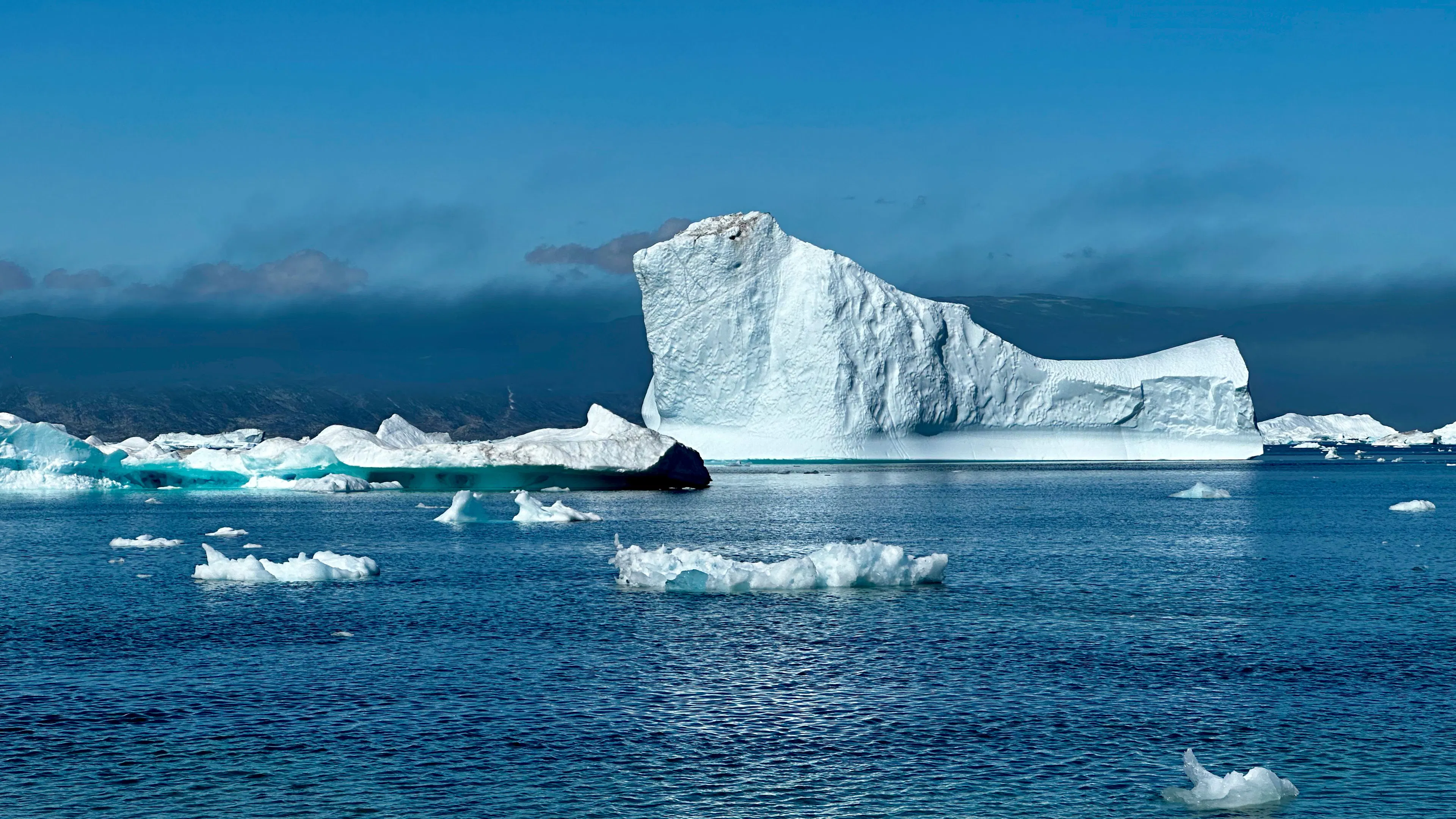 A tall white iceberg rising from deep blue Arctic water, with scattered ice floes in the foreground