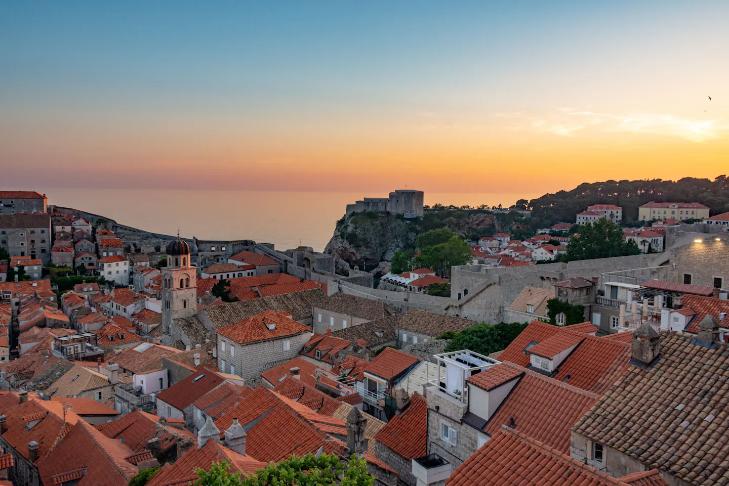 Dubrovnik's terracotta rooftops from the city walls at sunset, with the Franciscan bell tower, the sea beyond and Fort Lovrijenac on its cliff under an orange and blue sky
