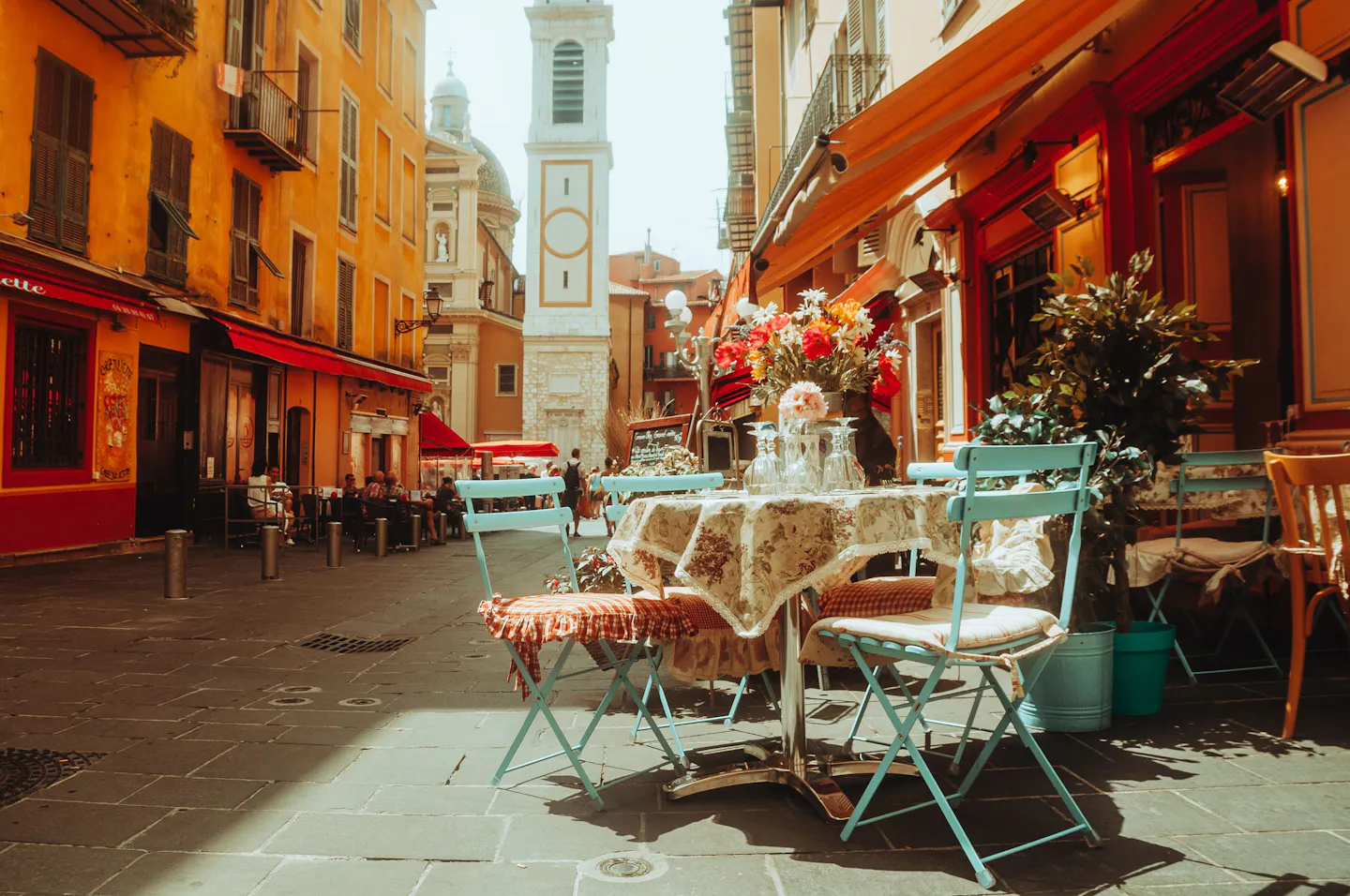 Café tables with flowers set out on a sunlit old-town street below a clock tower in Nice