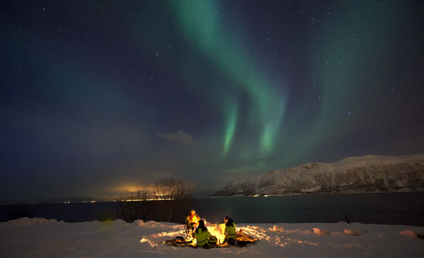 Three people around a campfire on a snowy shore watching green aurora over a fjord near Tromsø