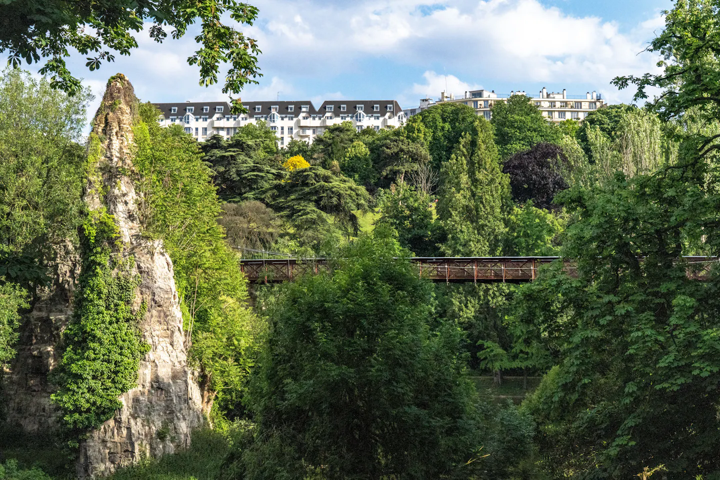 The ivy-covered rock pinnacle and red iron footbridge of the Parc des Buttes-Chaumont in Paris, surrounded by dense summer trees with Haussmann rooftops on the ridge above