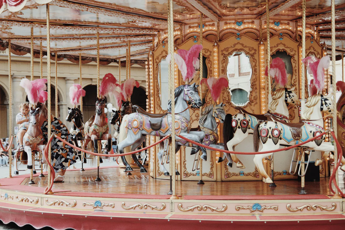 An ornate Belle Epoque carousel with gilded panels, bevelled mirrors and rows of painted horses wearing tall pink ostrich plumes, standing in front of a classical arcaded building