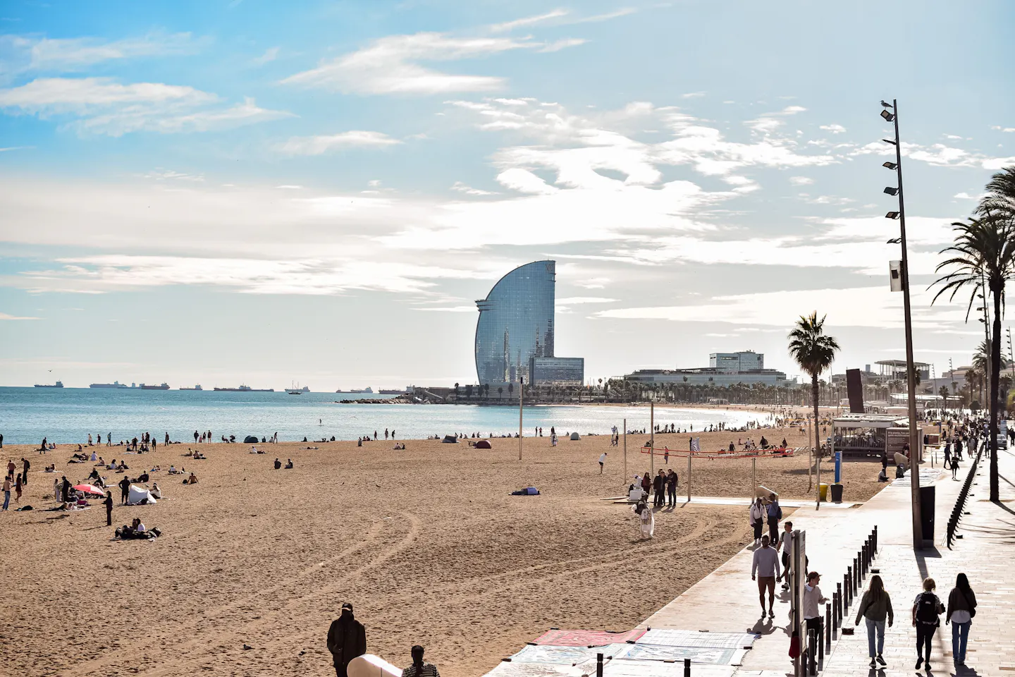 Barceloneta beach and its palm-lined promenade in afternoon light, with the sail-shaped hotel on the headland beyond