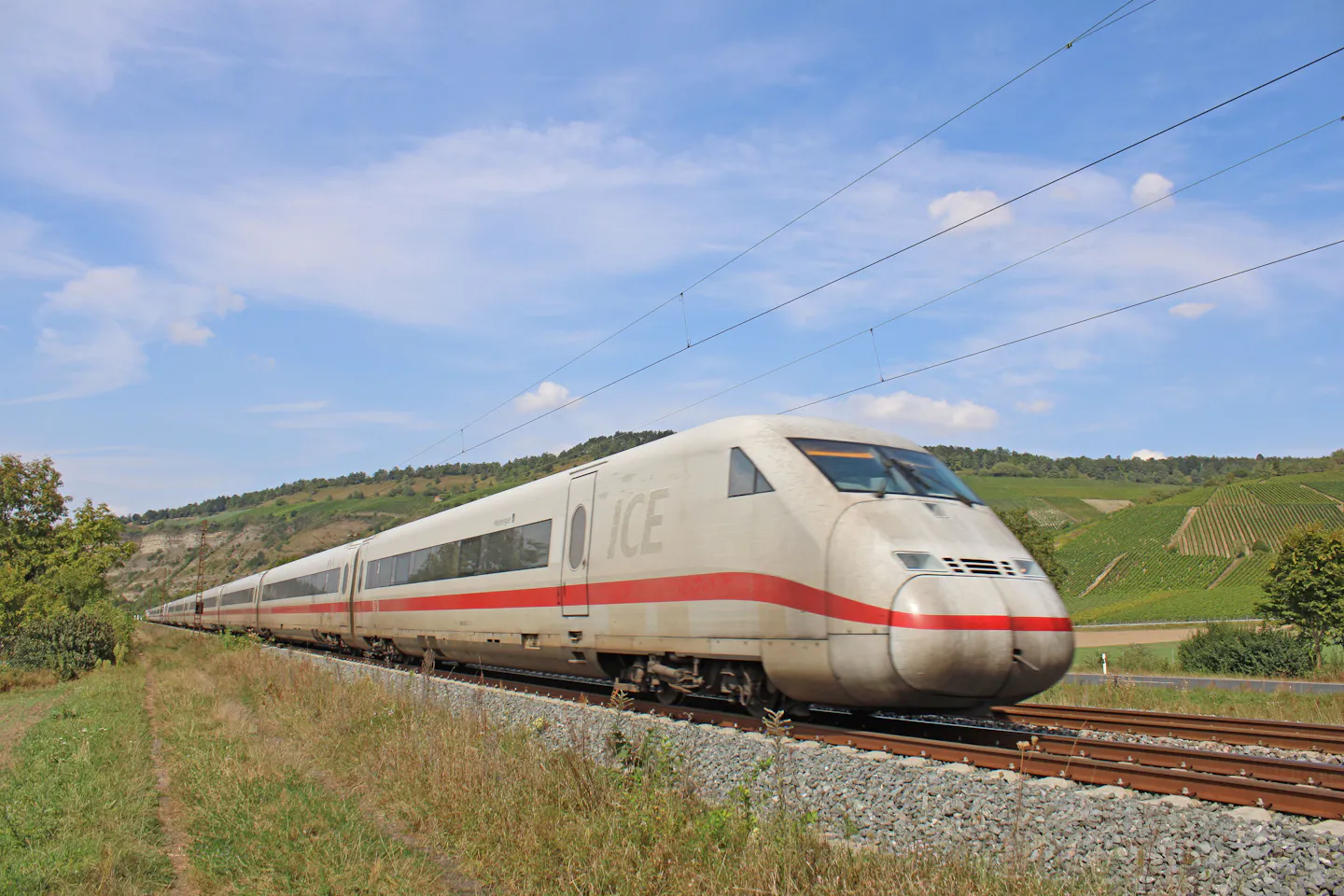A white and red high-speed train running past terraced vineyards under a blue sky