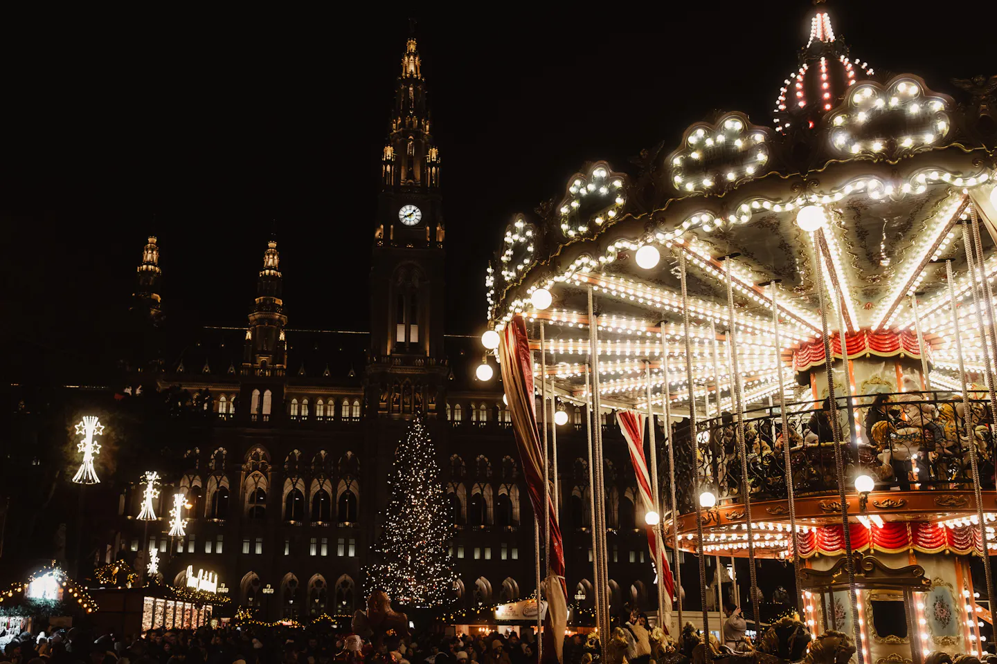 A lit carousel and Christmas tree at Vienna's Rathausplatz market with the City Hall tower behind