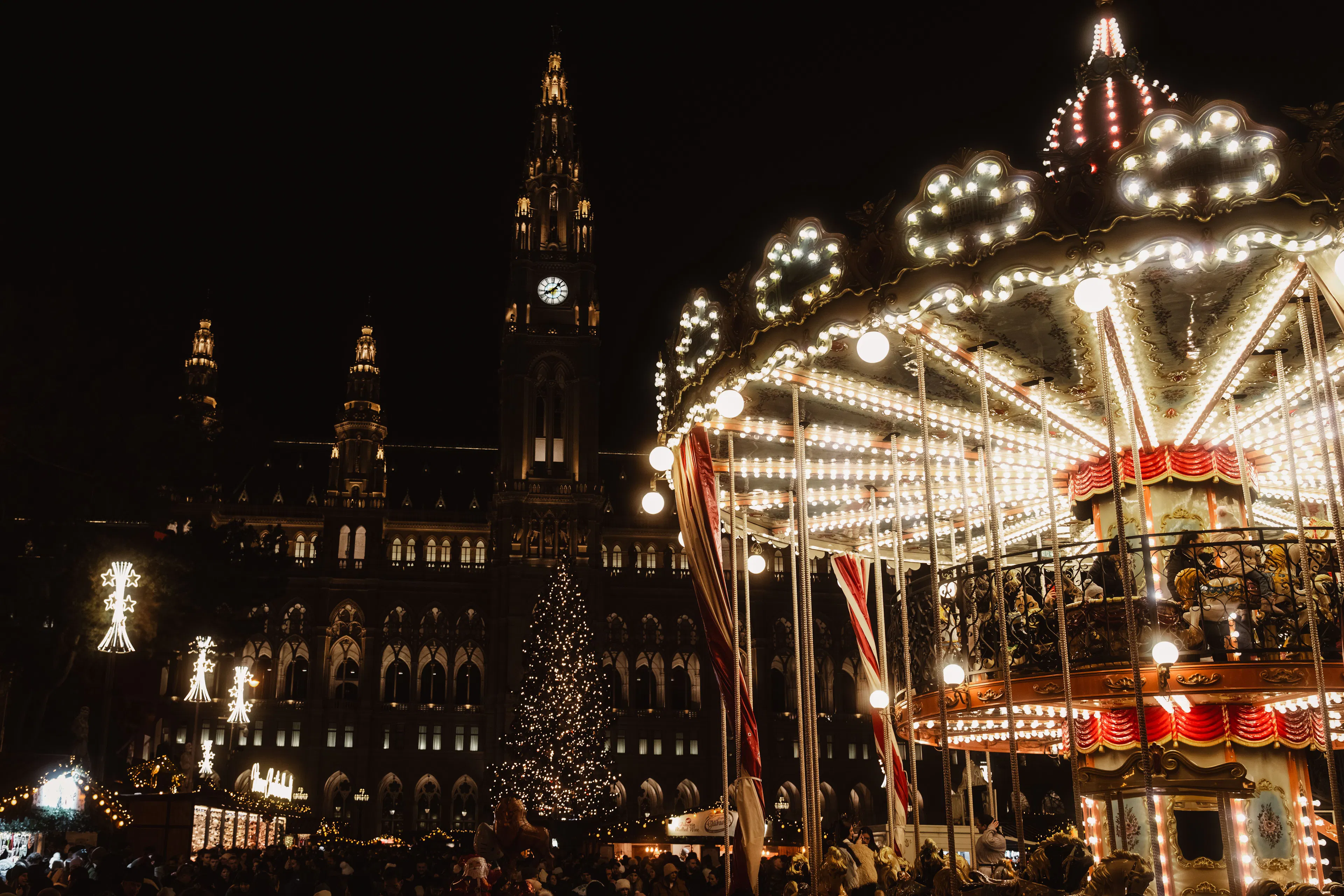 A lit carousel and Christmas tree at Vienna's Rathausplatz market with the City Hall tower behind