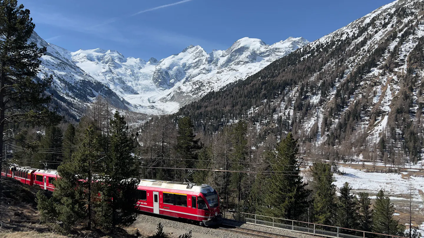 A red Alpine train curving through pine woods below glaciers and snow peaks in the Swiss Alps