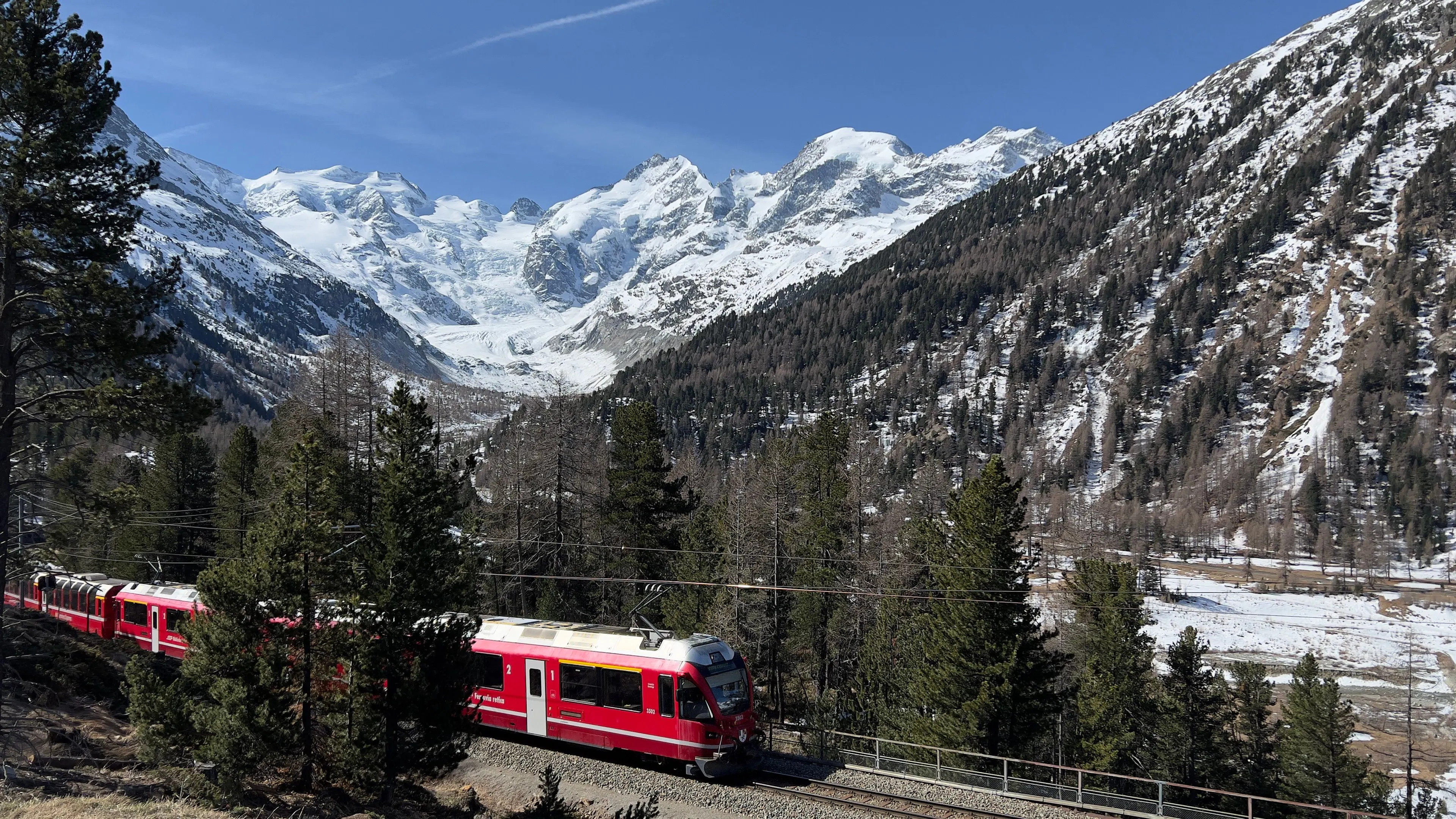 A red Alpine train curving through pine woods below glaciers and snow peaks in the Swiss Alps