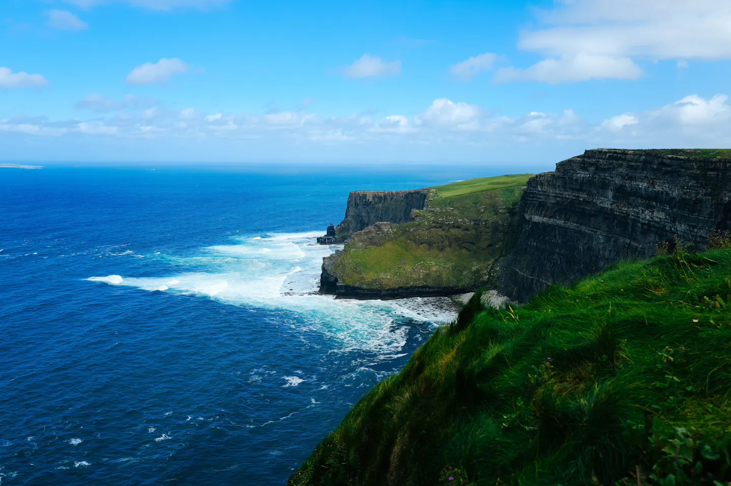 The Cliffs of Moher dropping to deep blue Atlantic water and white surf, with vivid green grass along the clifftop