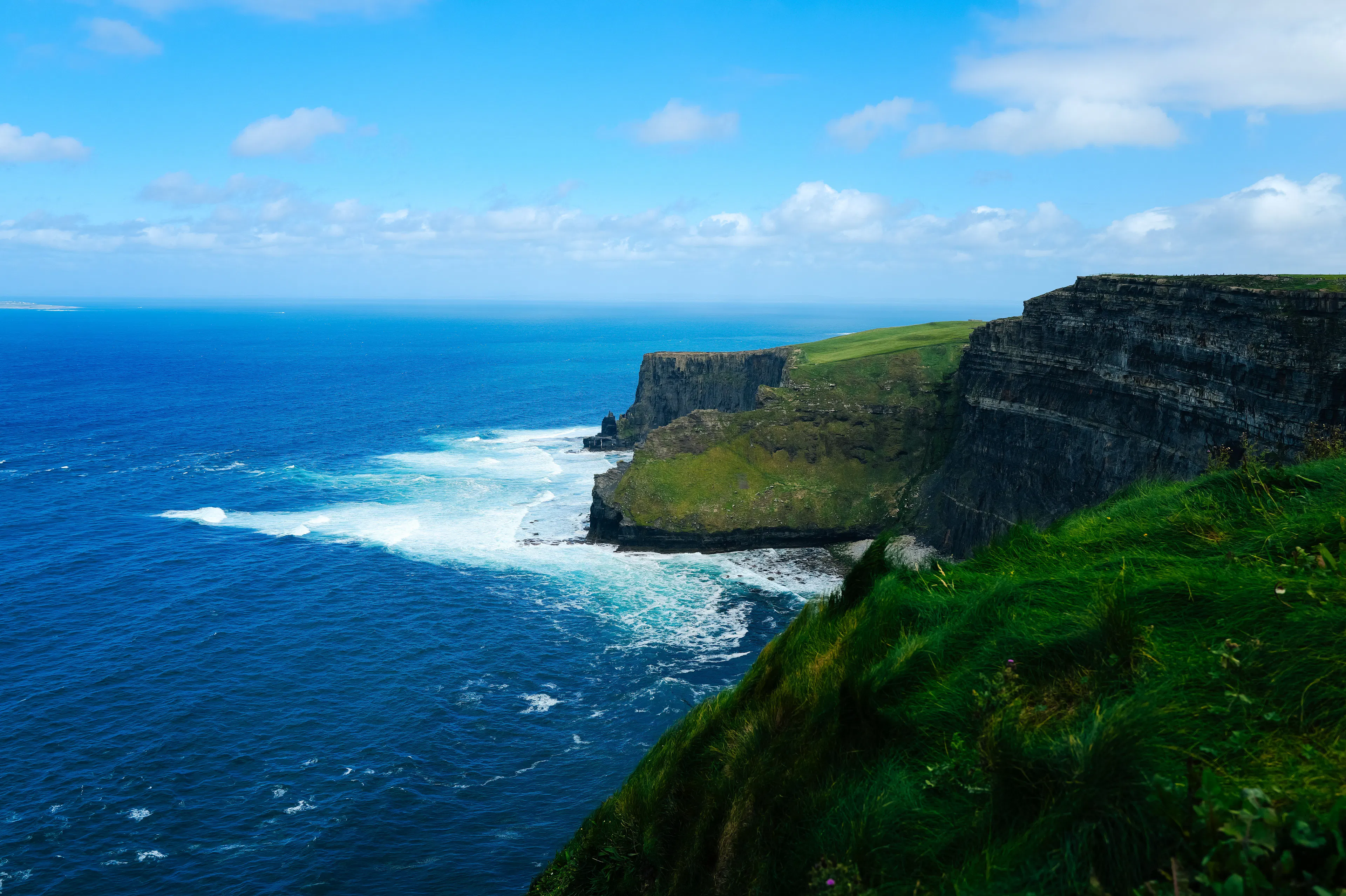 The Cliffs of Moher dropping to deep blue Atlantic water and white surf, with vivid green grass along the clifftop