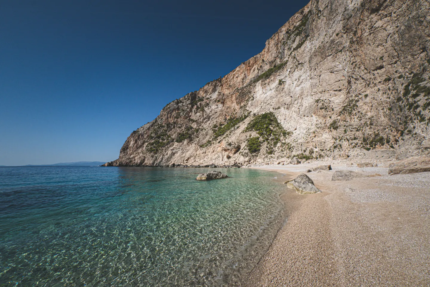 Empty pale pebble cove curving beneath a high limestone cliff, with clear turquoise shallows under a deep blue sky