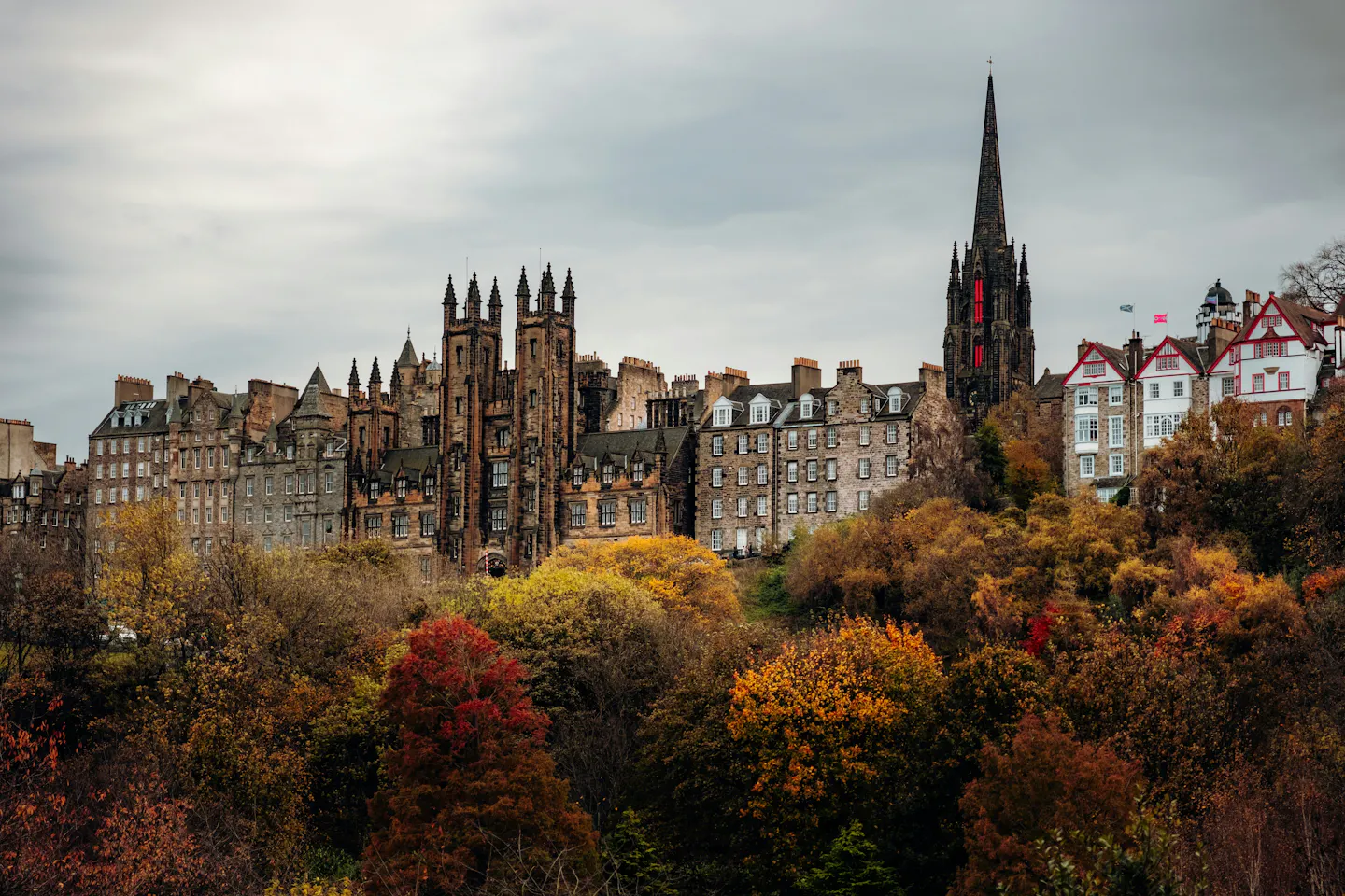 Edinburgh's Old Town skyline in autumn — the gothic towers of New College, the Tolbooth spire and the red-and-white gables of Ramsay Garden rising above a bank of rust, gold and scarlet trees