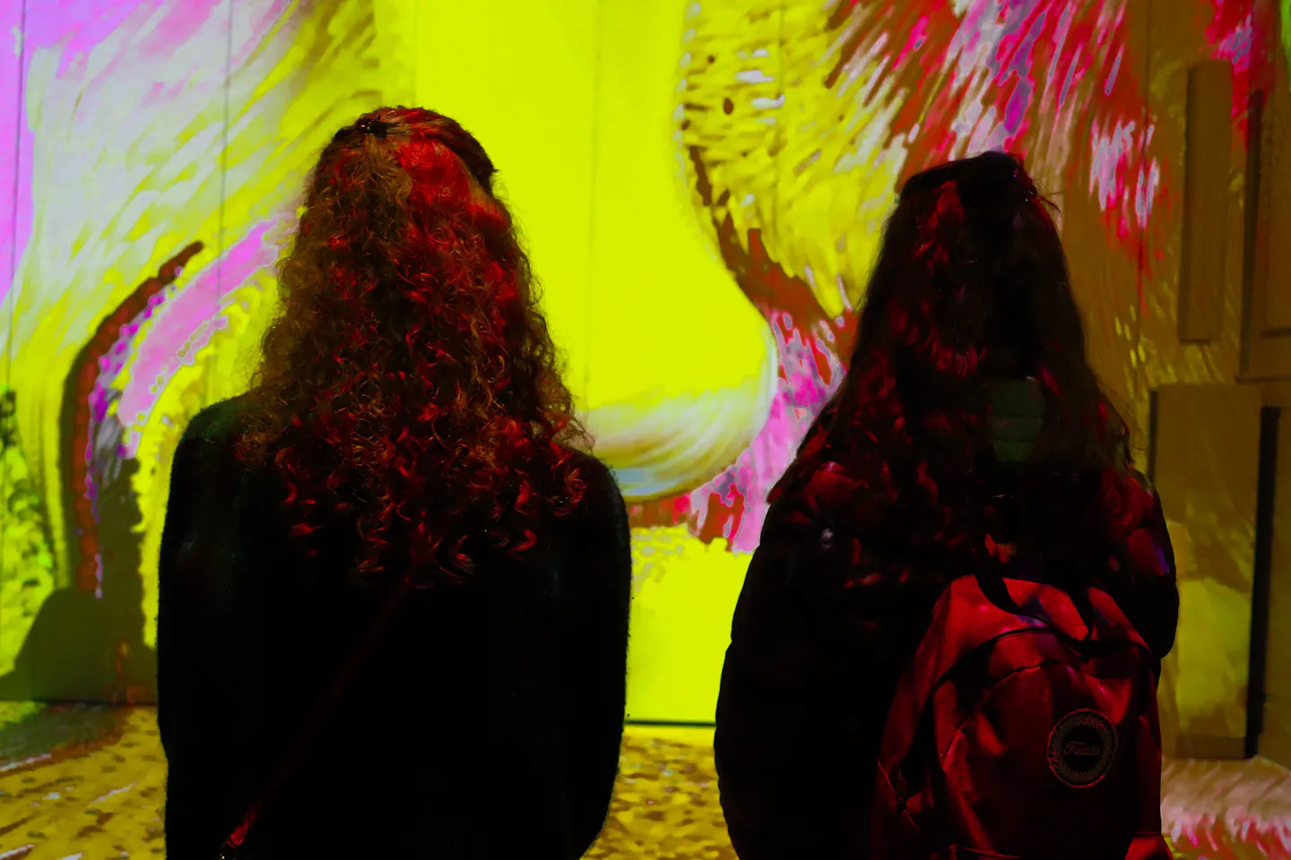 Two visitors silhouetted against a wall-sized projection of swirling yellow and pink brushstrokes in a darkened immersive-art hall
