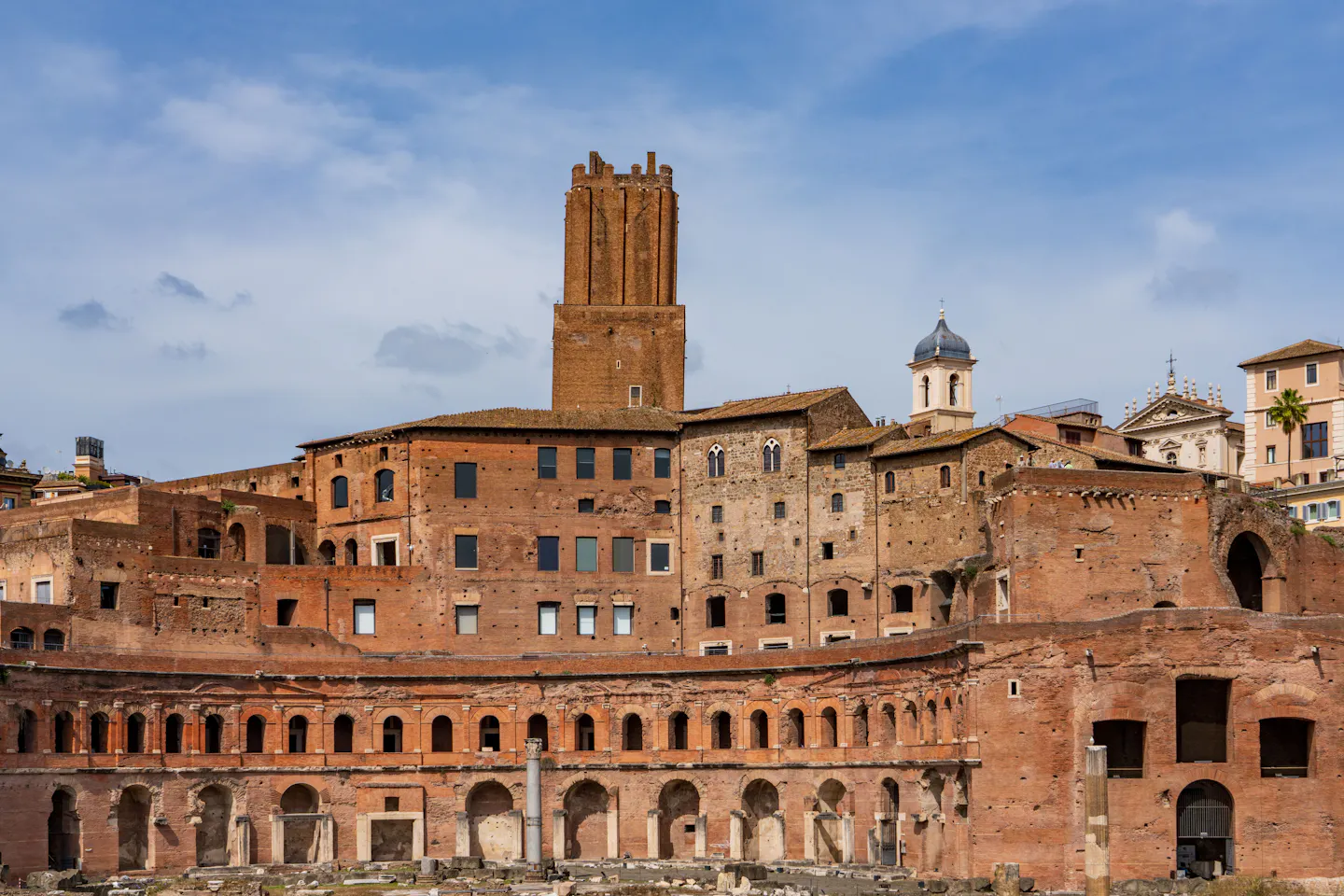 The tiered red-brick hemicycle of Trajan's Markets in Rome with its arched shopfronts and the medieval Torre delle Milizie above, under a blue sky with soft cloud