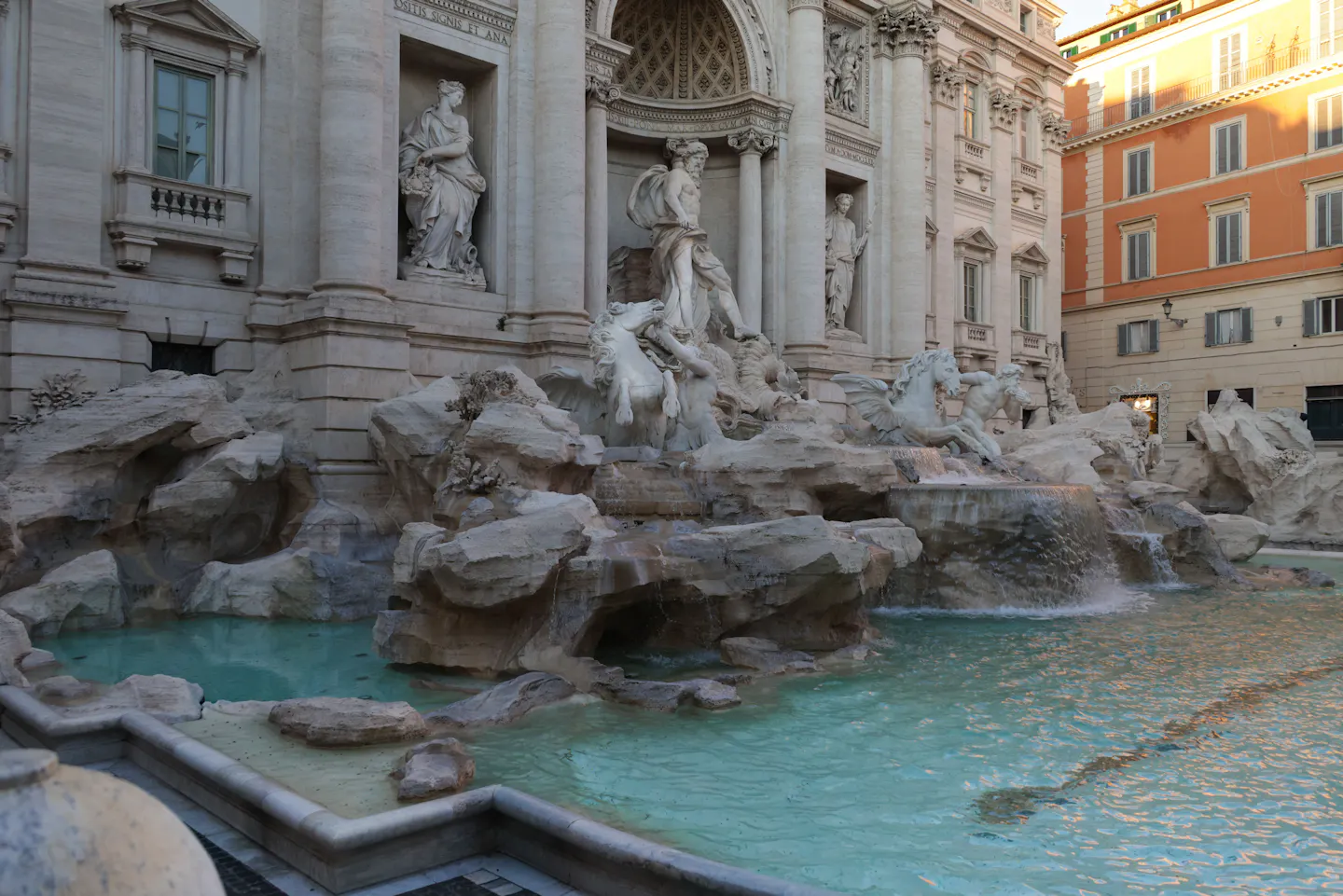 The Trevi Fountain early in the morning with nobody in front of it: Oceanus and the sea-horses on their travertine reef, water spilling into a still turquoise basin, warm sun catching the buildings on the right