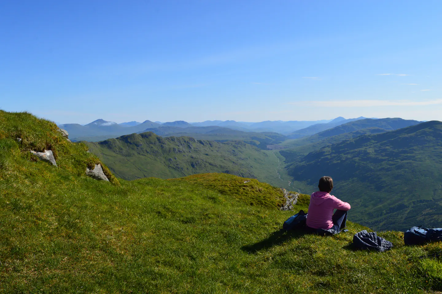 A hiker resting on a grassy Highland summit with jackets and a day bag, overlooking a green valley