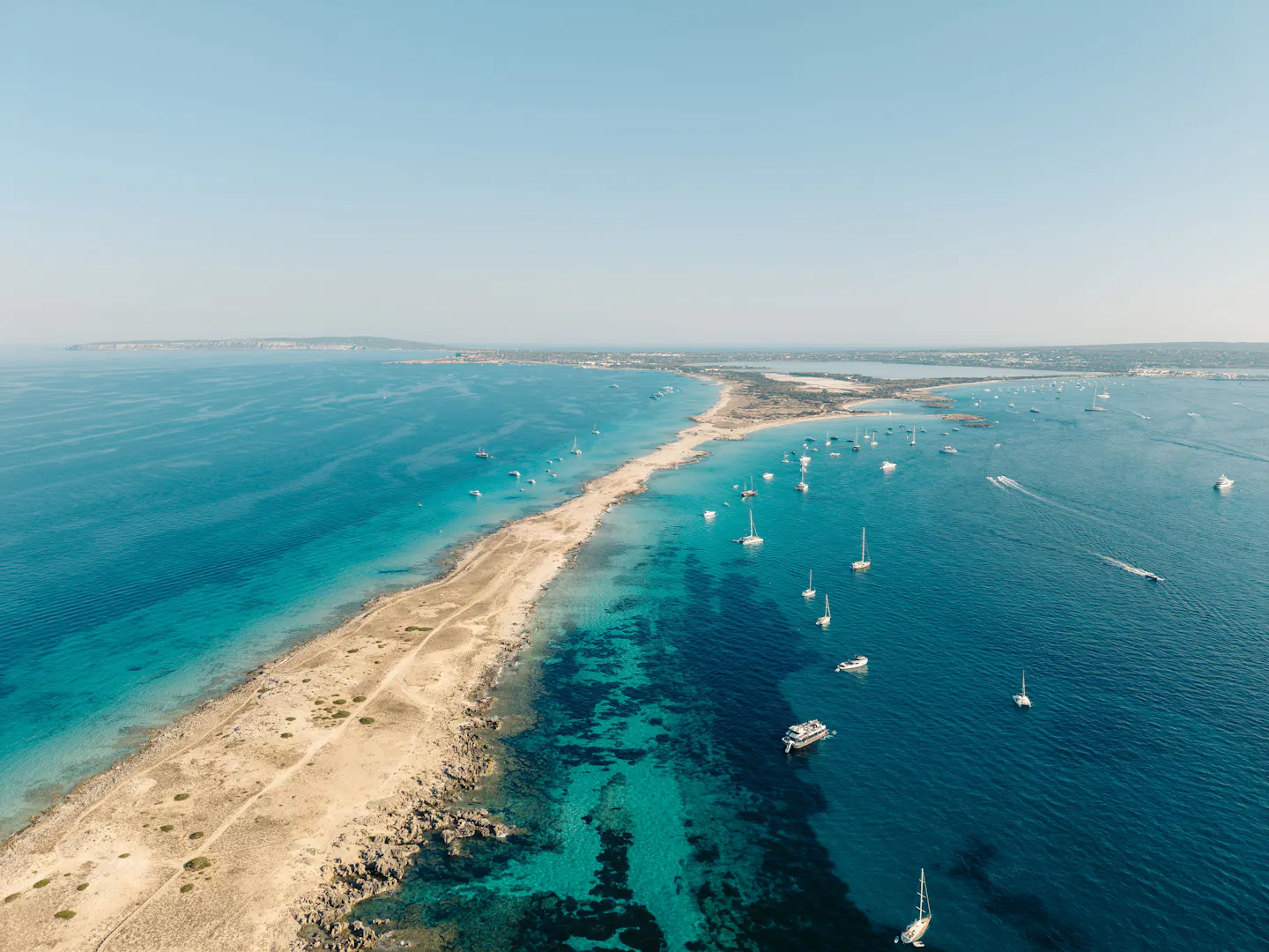 A narrow sandbar between turquoise shallows with dozens of yachts anchored off Formentera