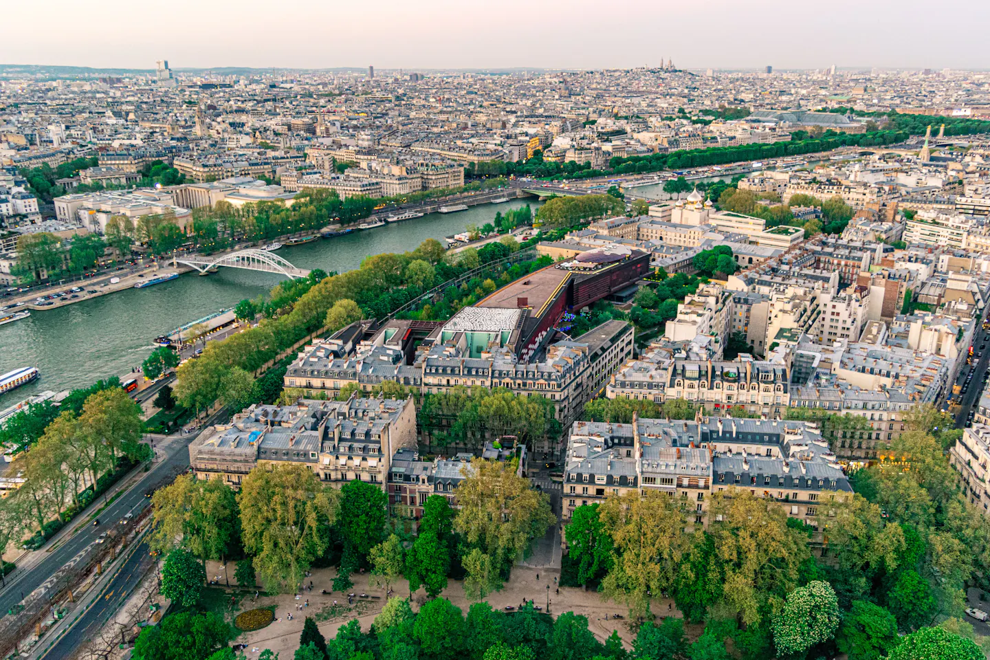 A high view over Paris at dusk: the Seine curving past a footbridge and museum roofs, dense zinc rooftops beyond, and Sacre-Coeur on the far horizon in pale pink light