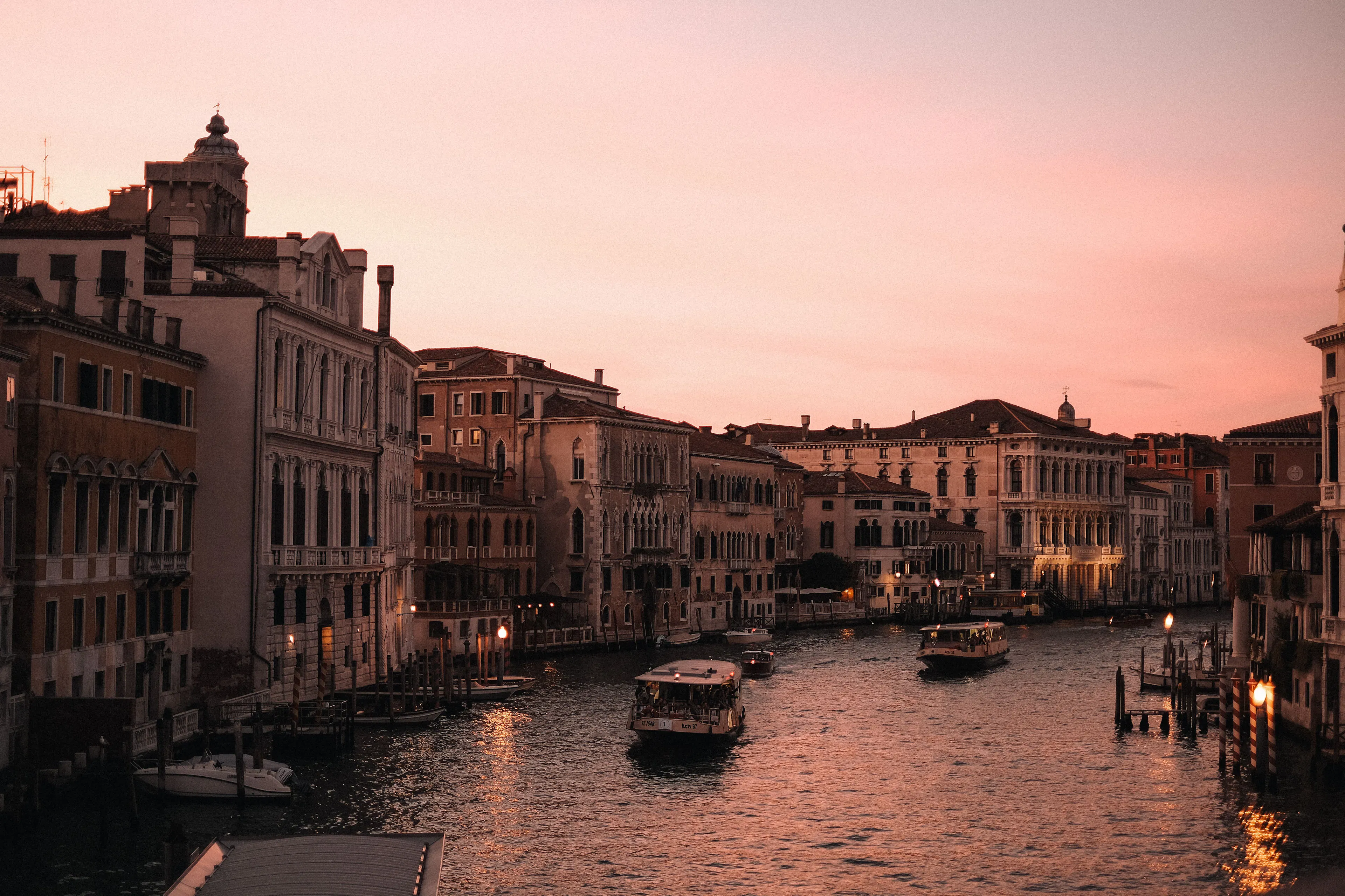 A wide Venetian canal at sunset, vaporetti passing lamplit palazzi under a pink sky