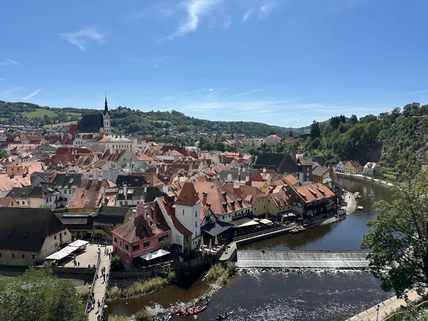 Český Krumlov from above in summer: red-tiled roofs and church spire of the old town with the Vltava curling around them past a weir and riverside terraces, under a bright blue sky