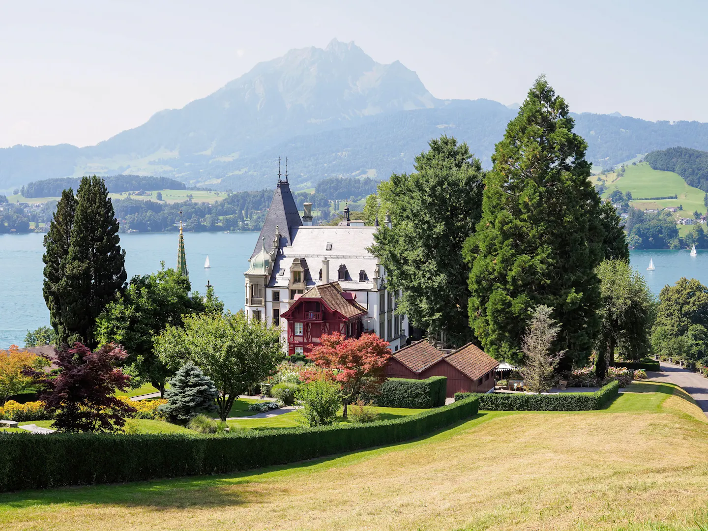 A lakeside chateau and its terraced gardens above Lake Lucerne, with sailboats on the water and Mount Pilatus rising behind
