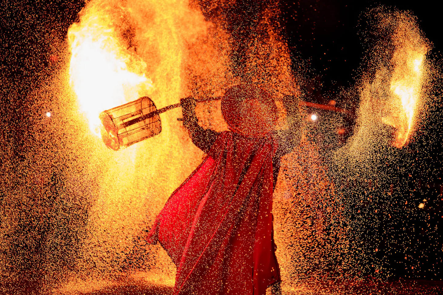 A robed performer in a deep red cloak swinging a burning steel cage at night, throwing a dense shower of orange sparks across the dark