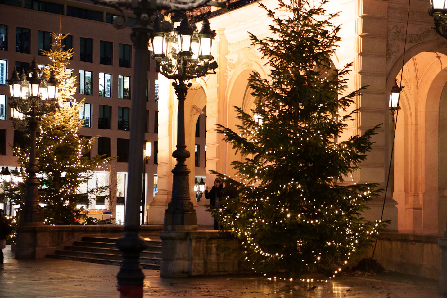 Two Christmas trees strung with warm lights beside ornate street lamps outside a stone arcade