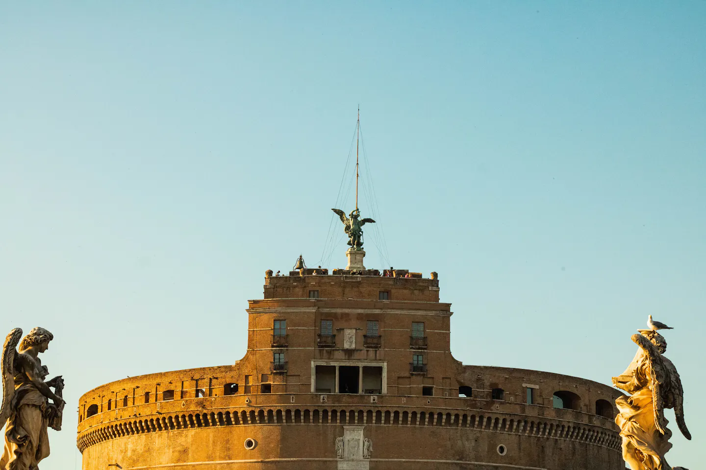 The bronze archangel on top of Castel Sant'Angelo in golden light, framed by two of the marble angel statues on Ponte Sant'Angelo