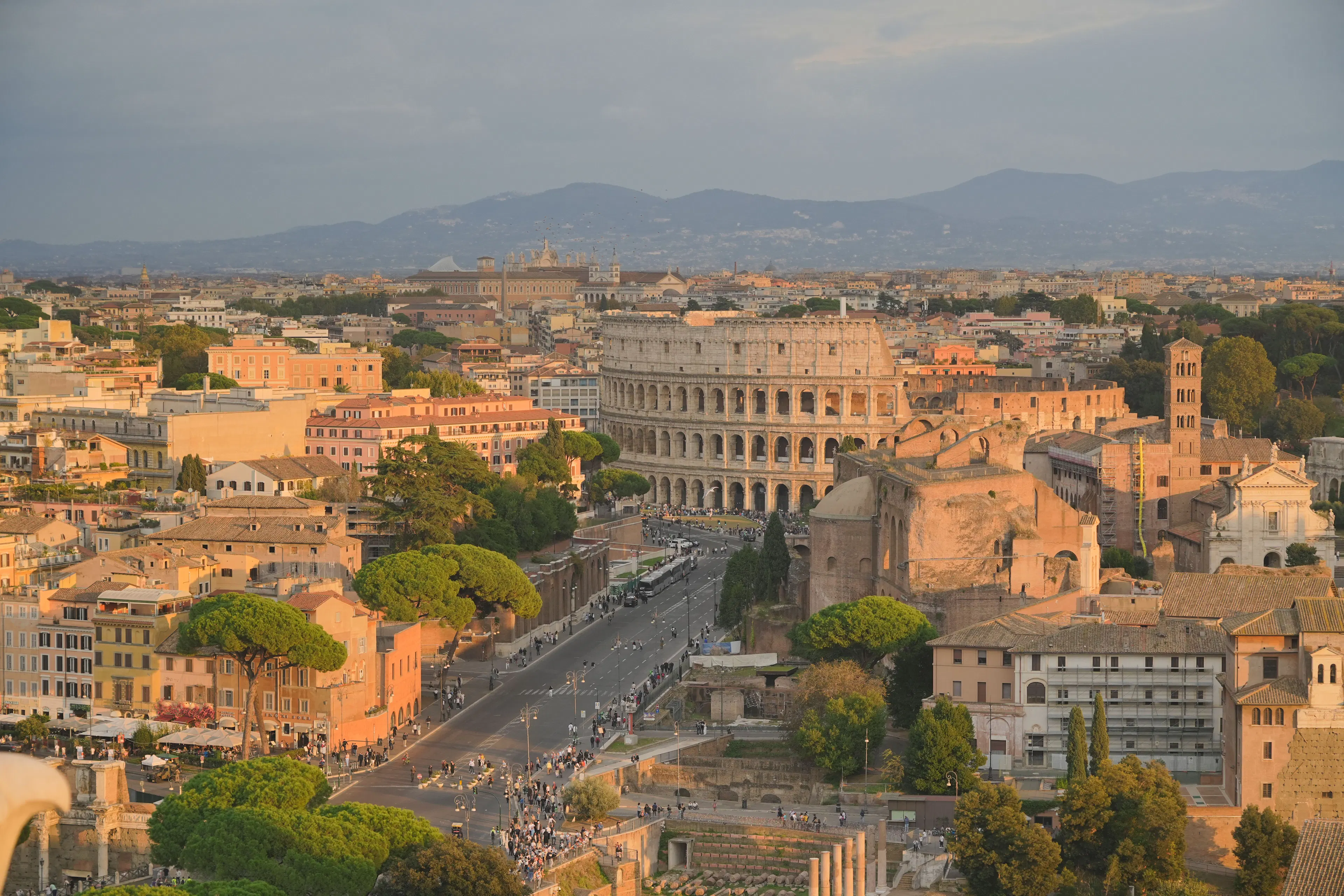 The Colosseum and the ruins of the Roman Forum seen from above in warm late-afternoon light, with umbrella pines and hills beyond the city