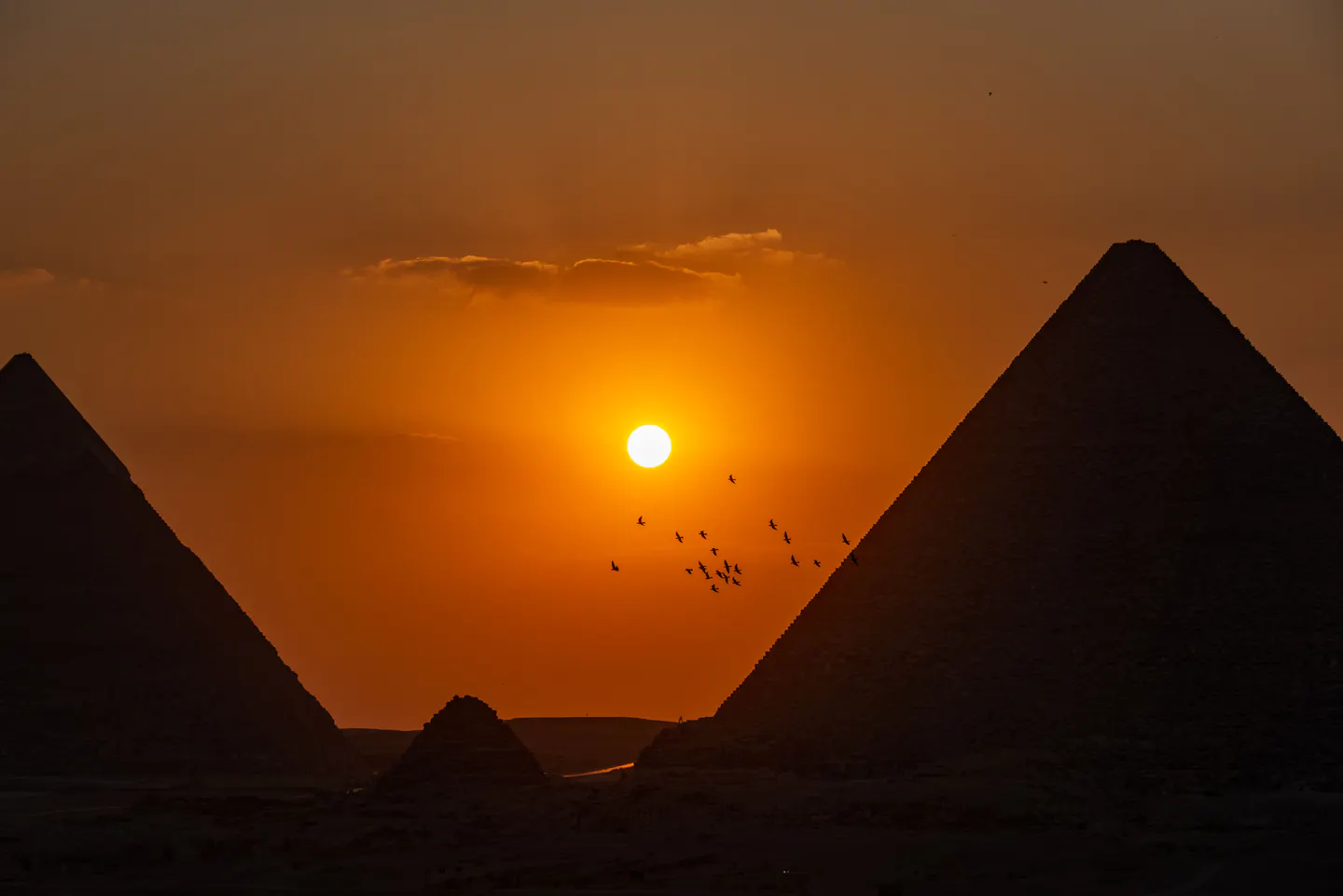 The Pyramids of Giza silhouetted against a deep orange sunset sky, the sun low in the gap between them and a flock of birds crossing the frame