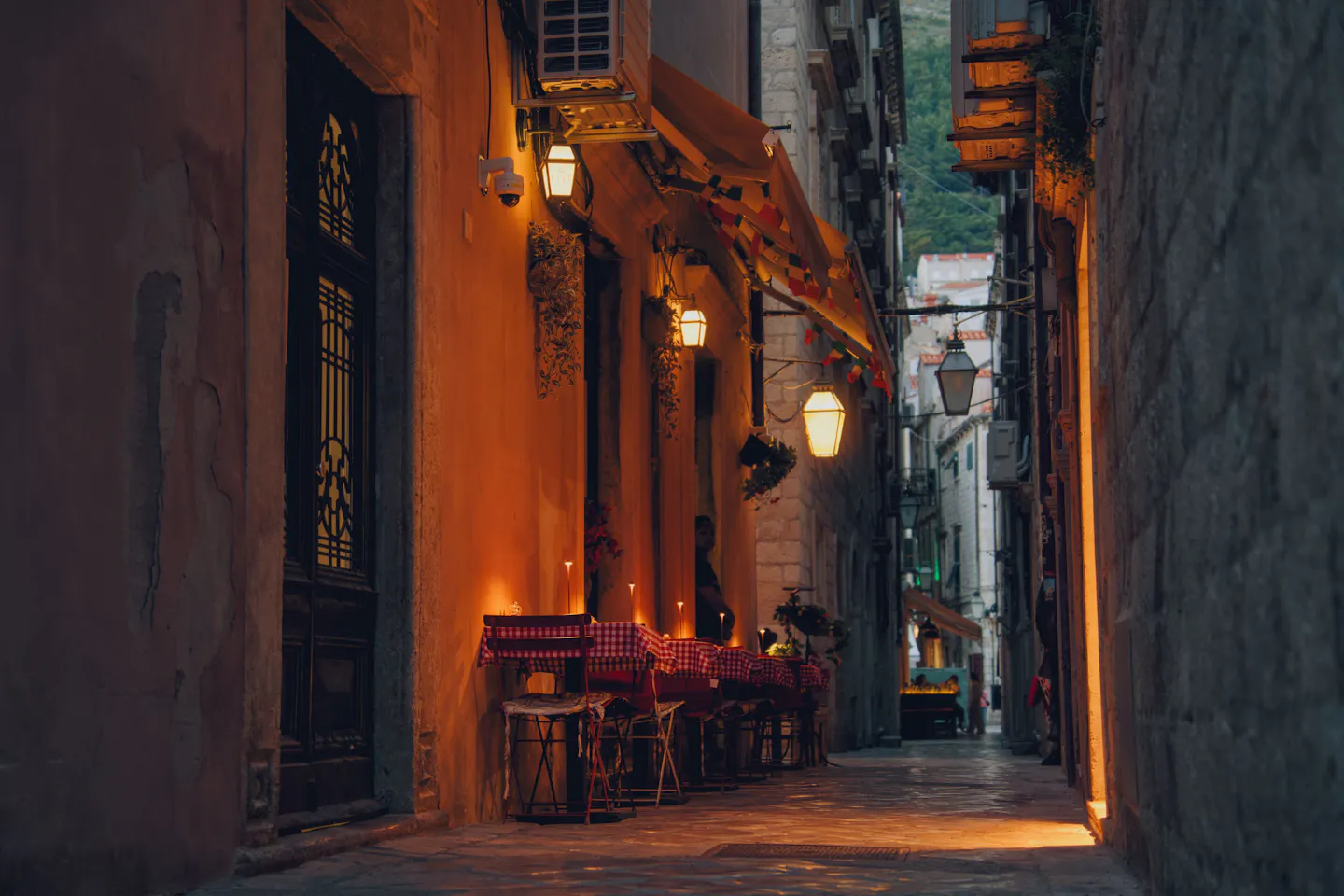 Warmly lit restaurant tables and hanging lanterns lining a narrow historic stone alley in the evening