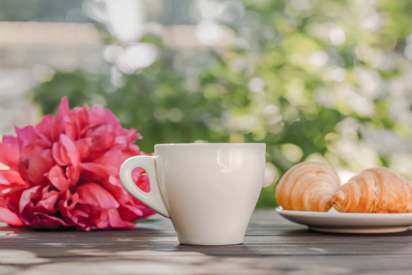 A white cup of coffee and two croissants on a wooden café table outdoors, beside a bright pink flower