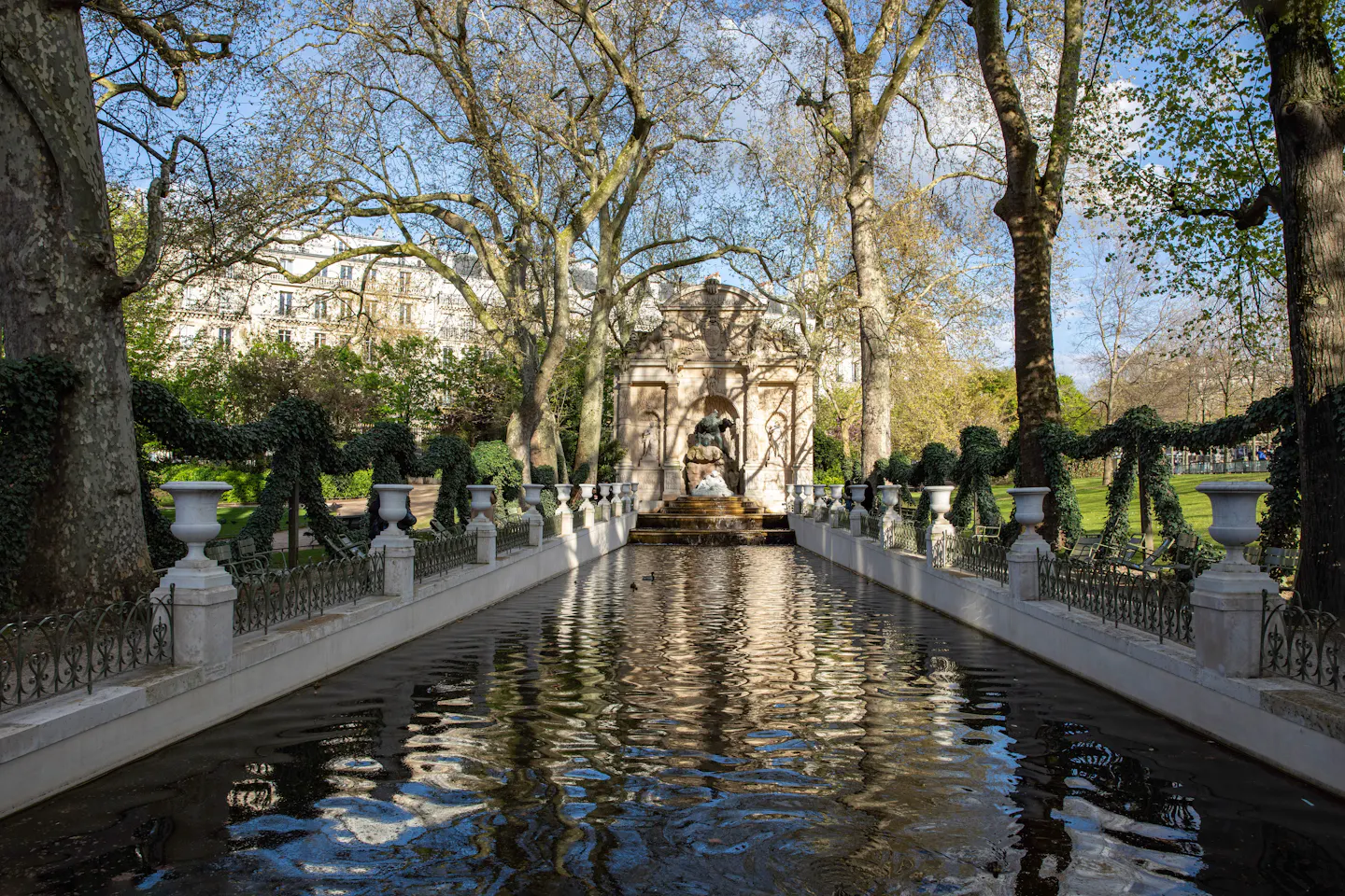 The Medici Fountain in the Luxembourg Garden reflected the length of its long rectangular basin, framed by plane trees, stone urns and ivy garlands in early spring sunlight