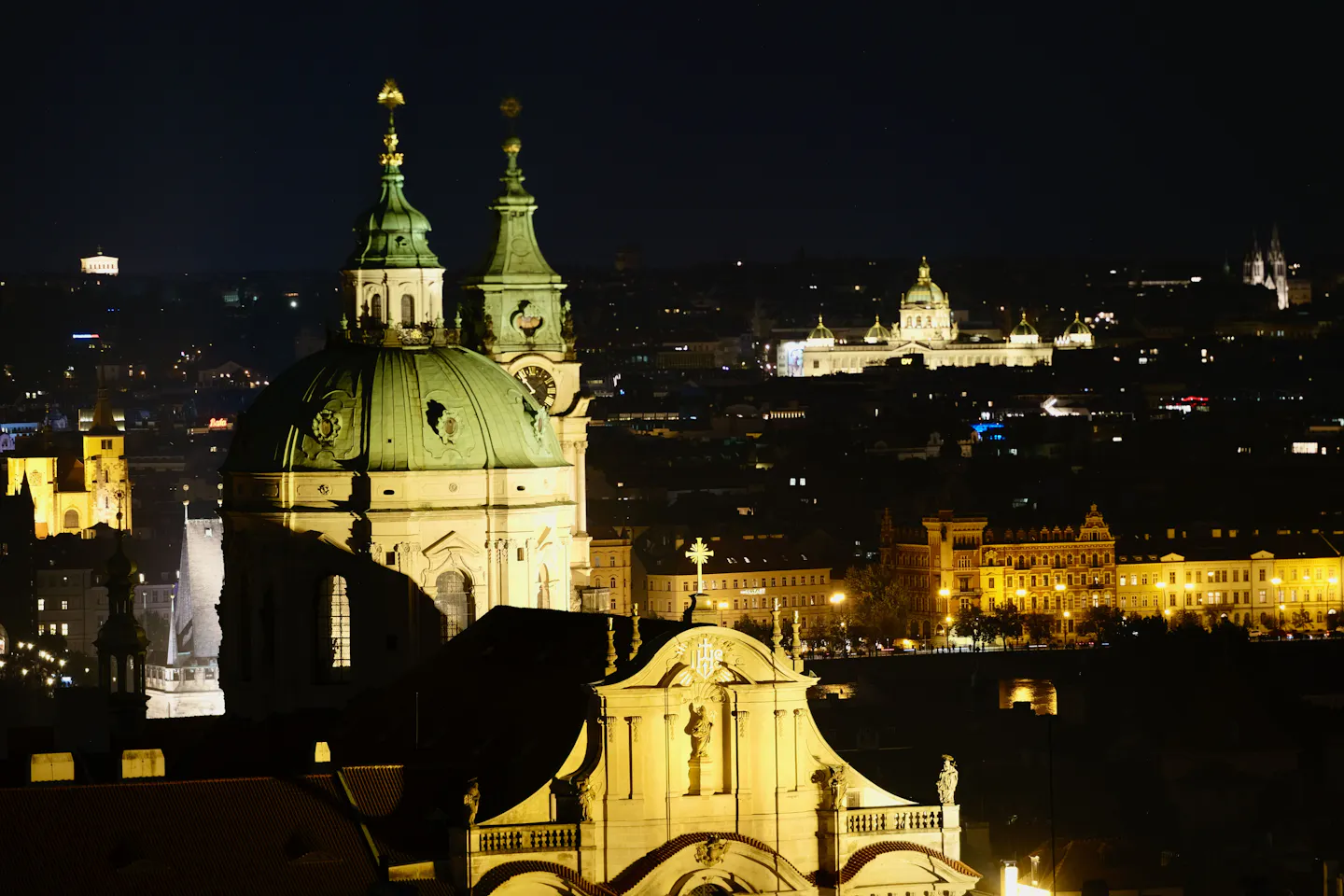 A floodlit baroque church dome and bell tower above Prague’s rooftops at night, with the lit city stretching away behind