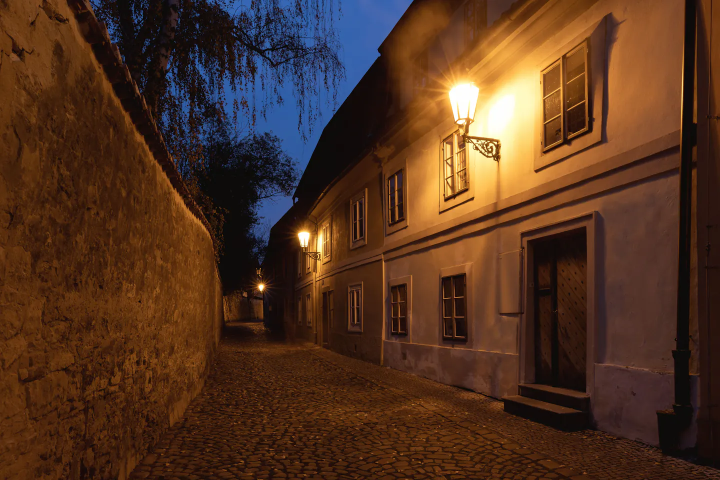 A cobbled Prague lane at blue hour lit by wrought-iron gas lamps on a pale stucco wall