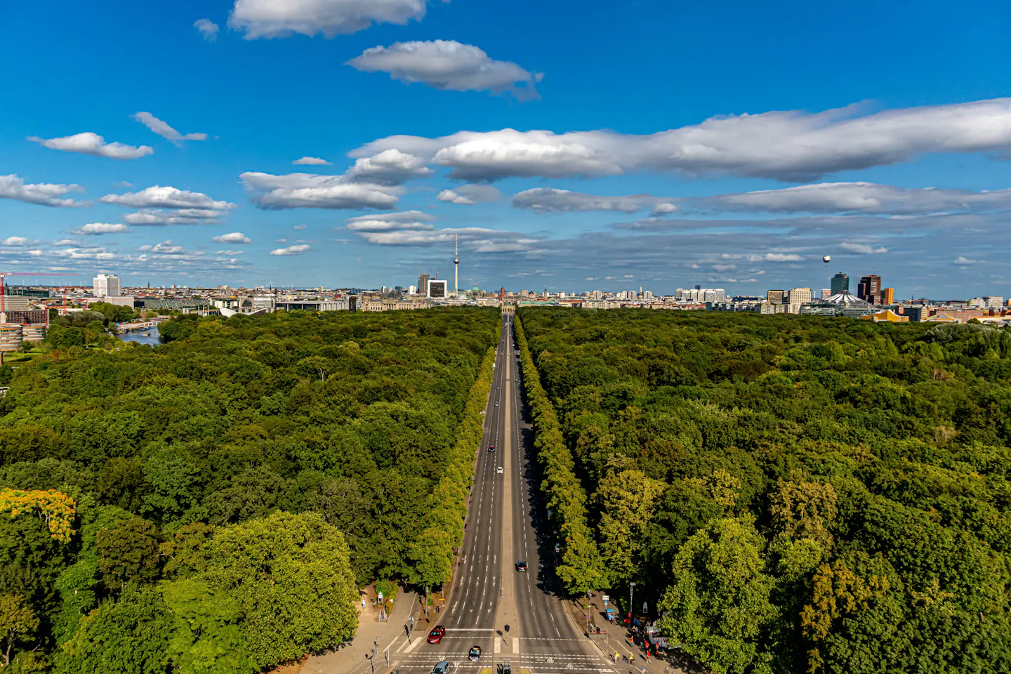 Aerial view over the dense green canopy of Berlin's Tiergarten, a straight avenue cutting through it, with the Fernsehturm and the city skyline on the horizon under a blue sky with cumulus cloud