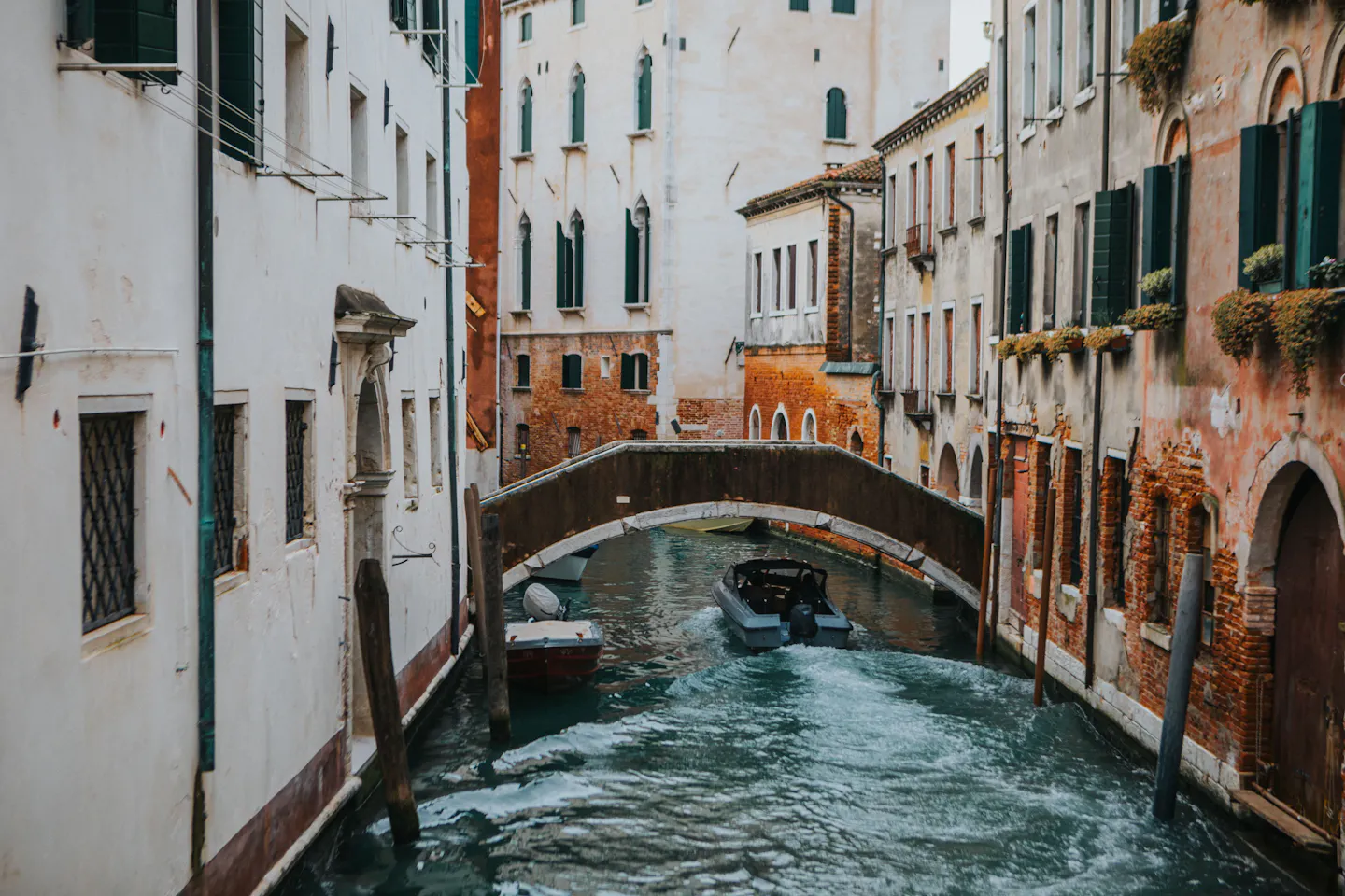 A small boat passing under a stone bridge on a narrow Venetian canal lined with brick and ivy