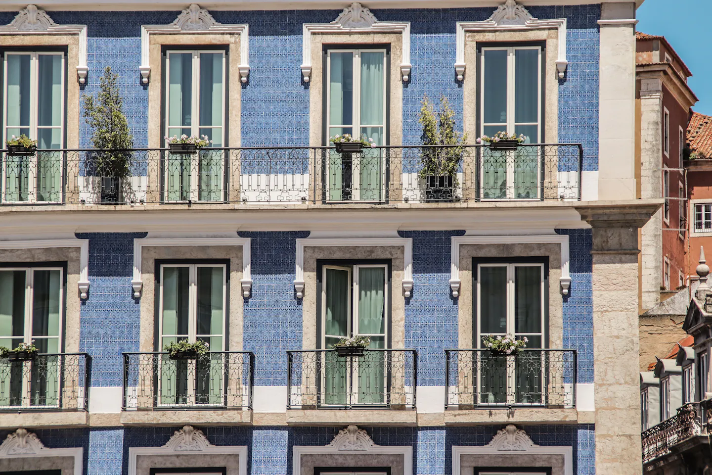 A Lisbon facade tiled entirely in blue-and-white azulejos, with wrought-iron balconies, green shutters and flower boxes lit by strong sun