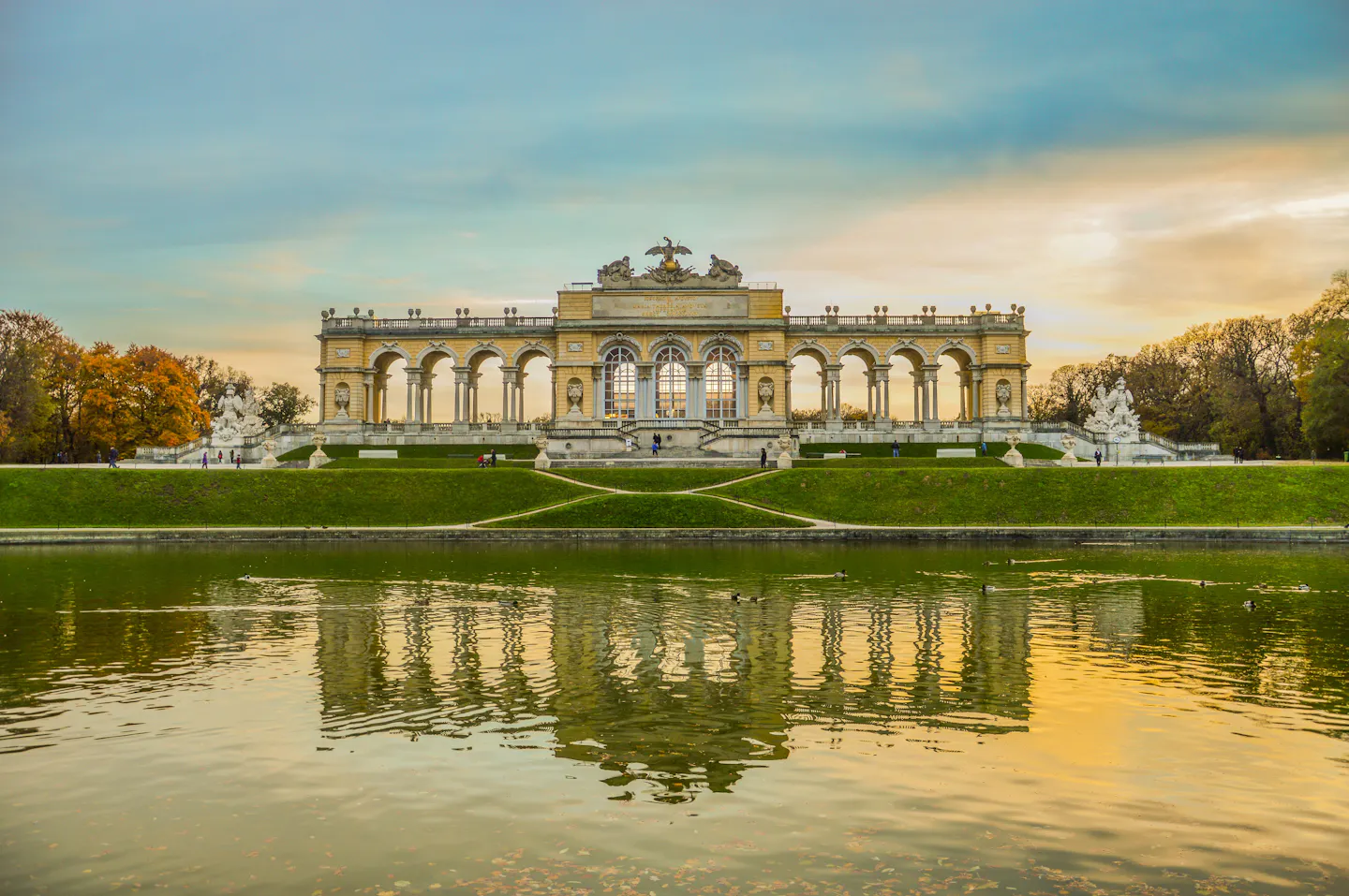 The Gloriette in Vienna's Schönbrunn gardens reflected in its pond at sunset, with autumn trees on either side