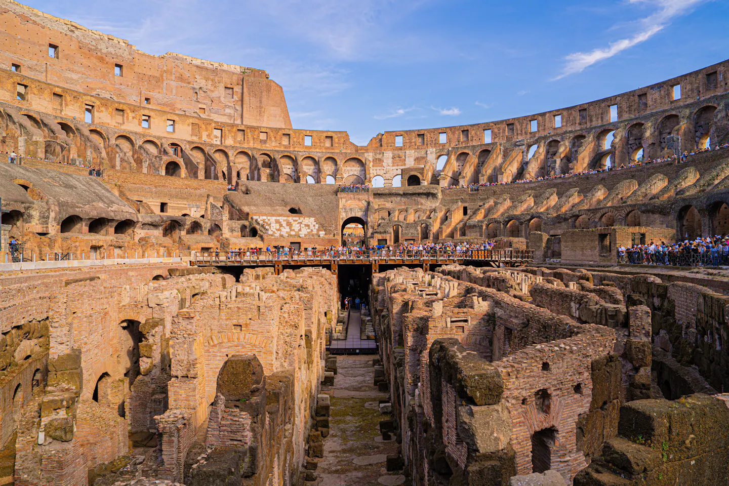 The Colosseum seen from the upper tiers, with the wooden arena walkway and the exposed underground passages below under a blue sky